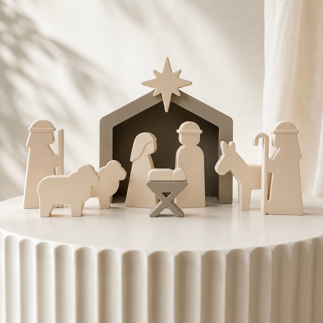 A minimalist silicone nativity set displayed on a white ribbed pedestal. The set includes a stable with a star, Mary, Joseph, baby Jesus in a manger, a shepherd, and an animal, all in a light natural wood finish with dark interior shadows.
