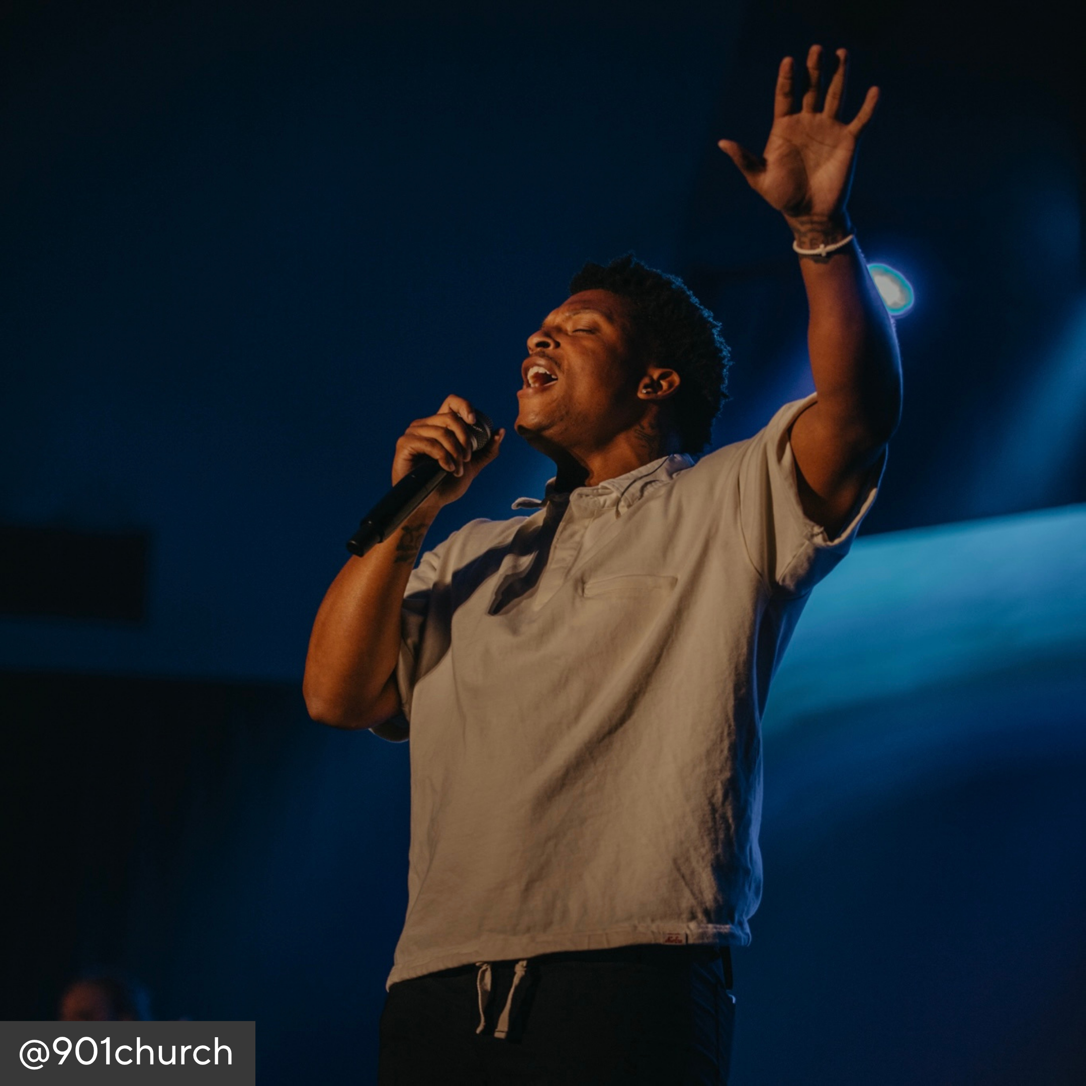 Person singing into a microphone with one hand raised, against a dark background.