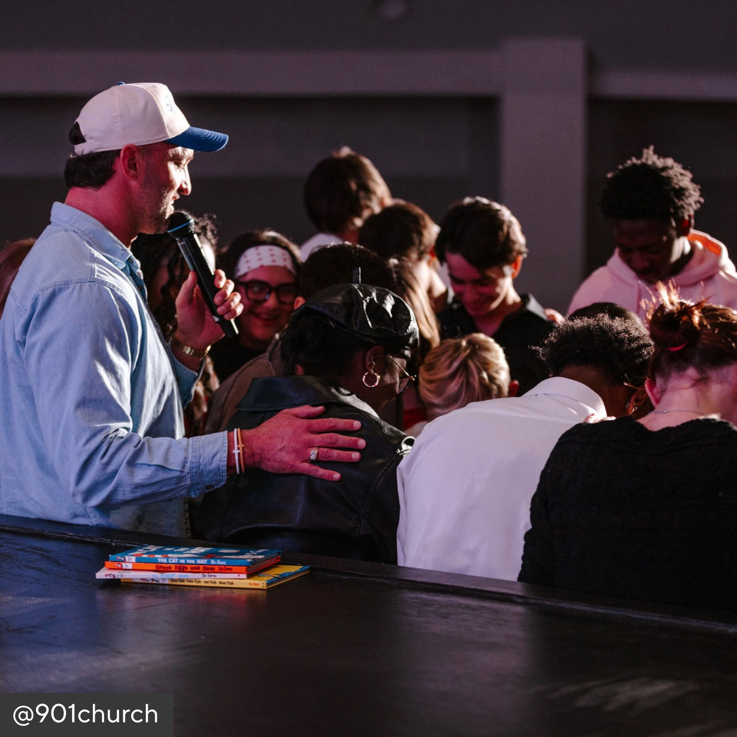 Group of people gathered around a man with a microphone, possibly in a church setting.