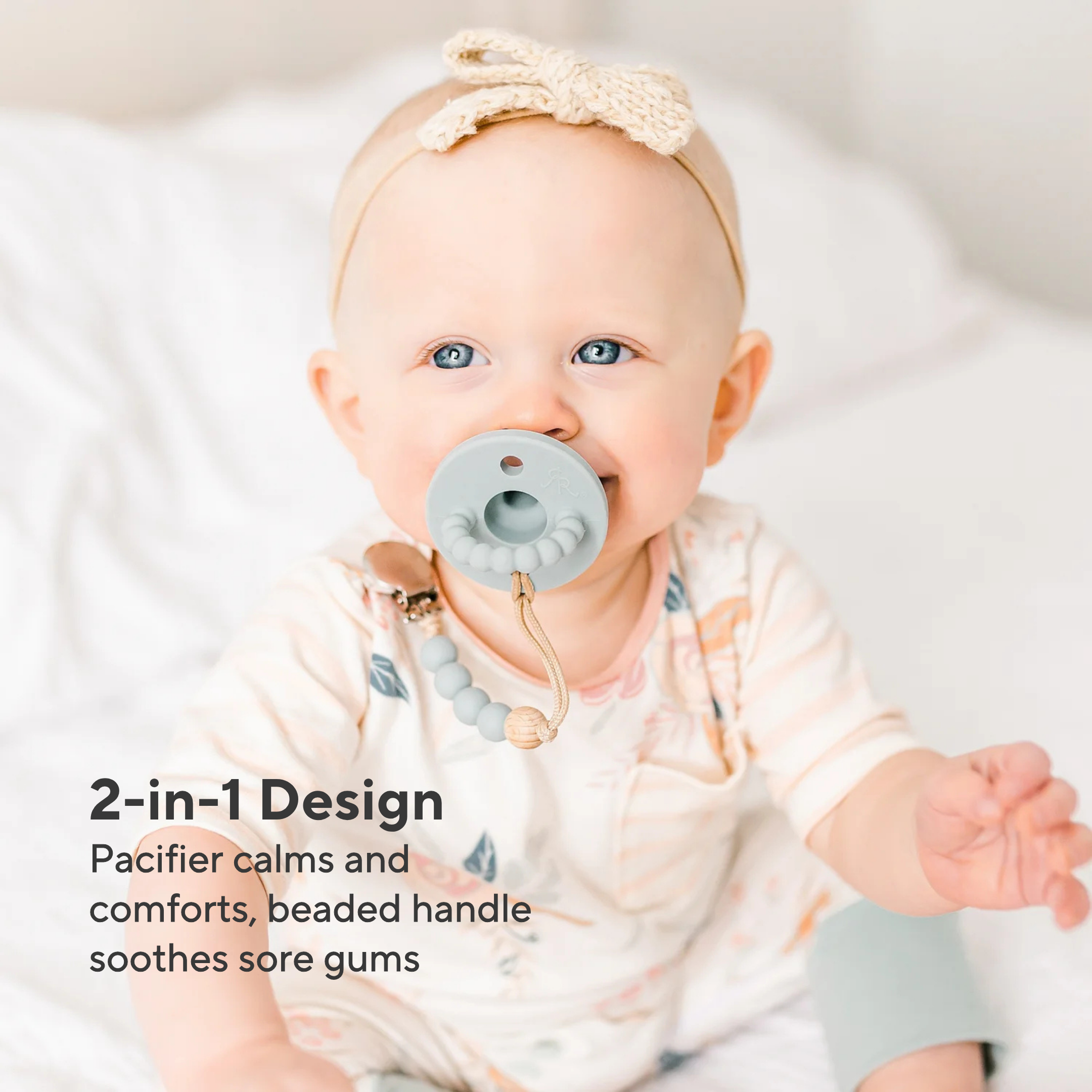 Baby wearing a pacifier with a beaded handle, sitting on a white blanket.