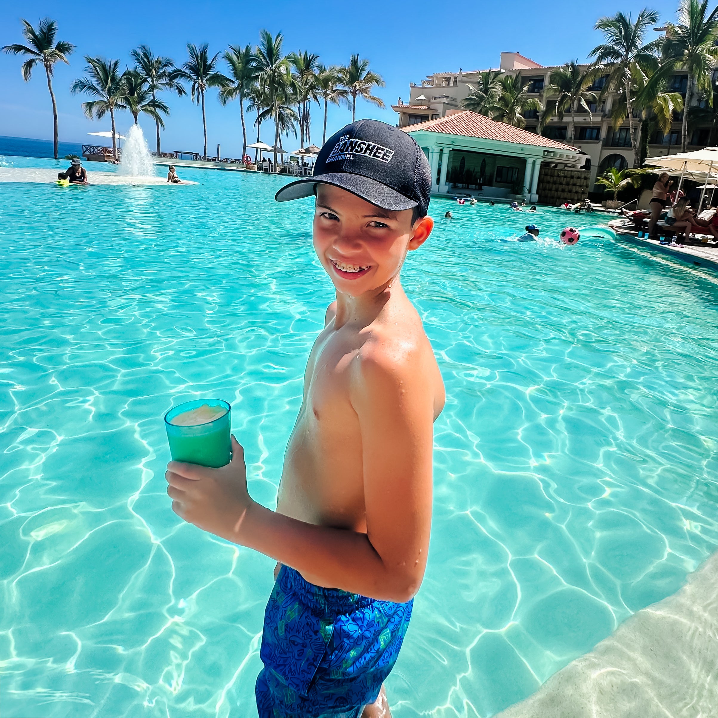 Young boy in a pool holding a cup with palm trees and a building in the background