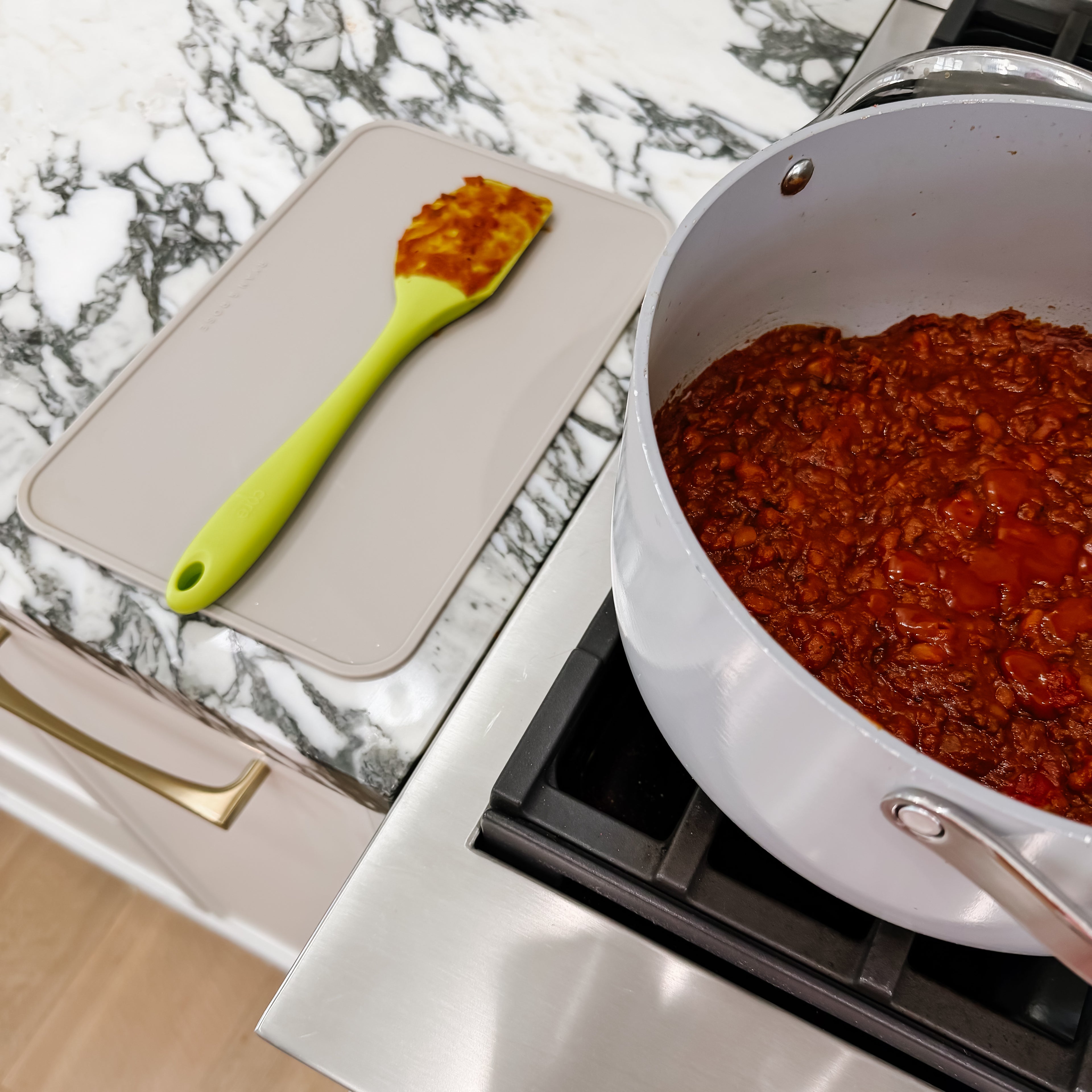 Pot of red sauce on a stove with a green spatula on a marble countertop.