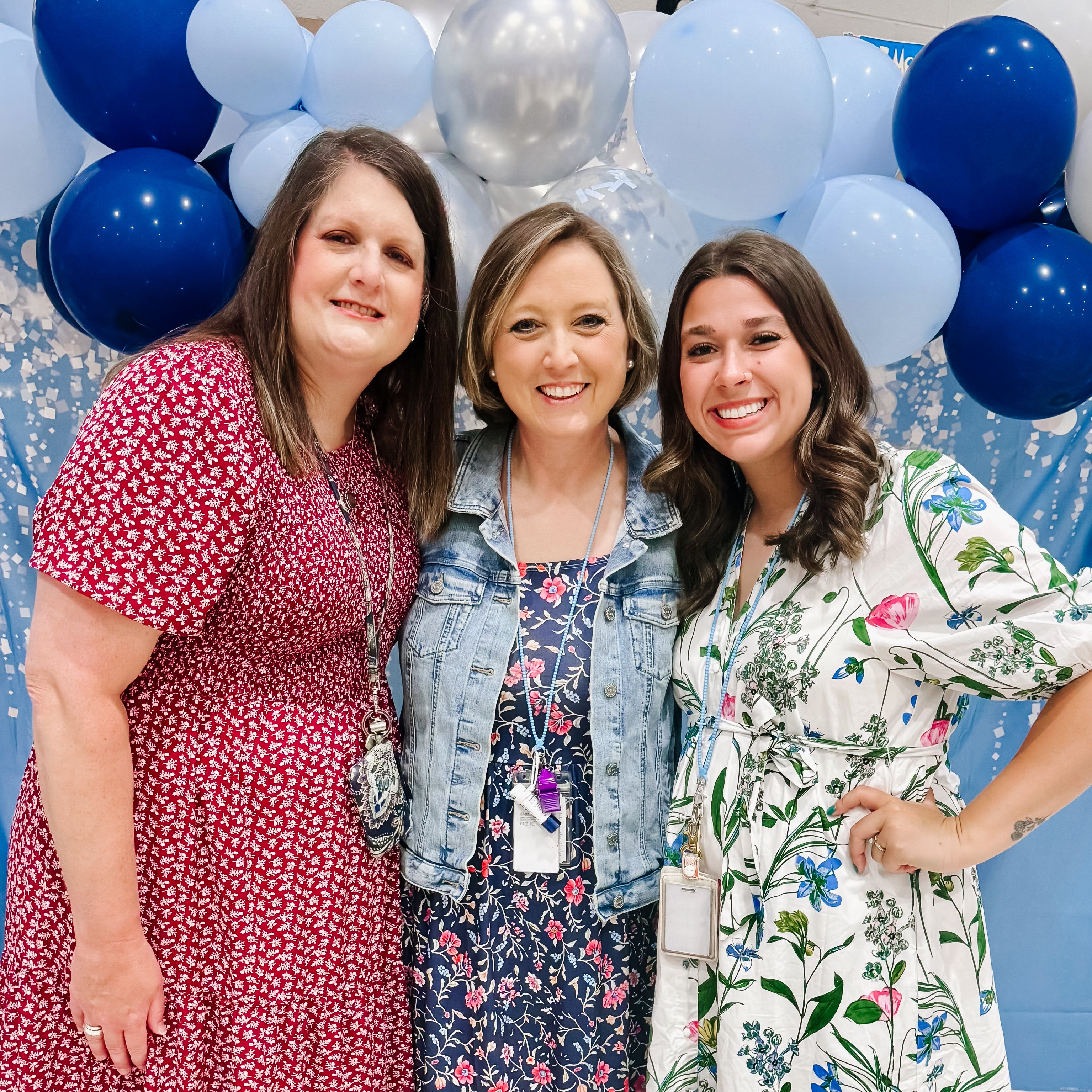 Three women posing together with blue, silver, and white balloons in the background.