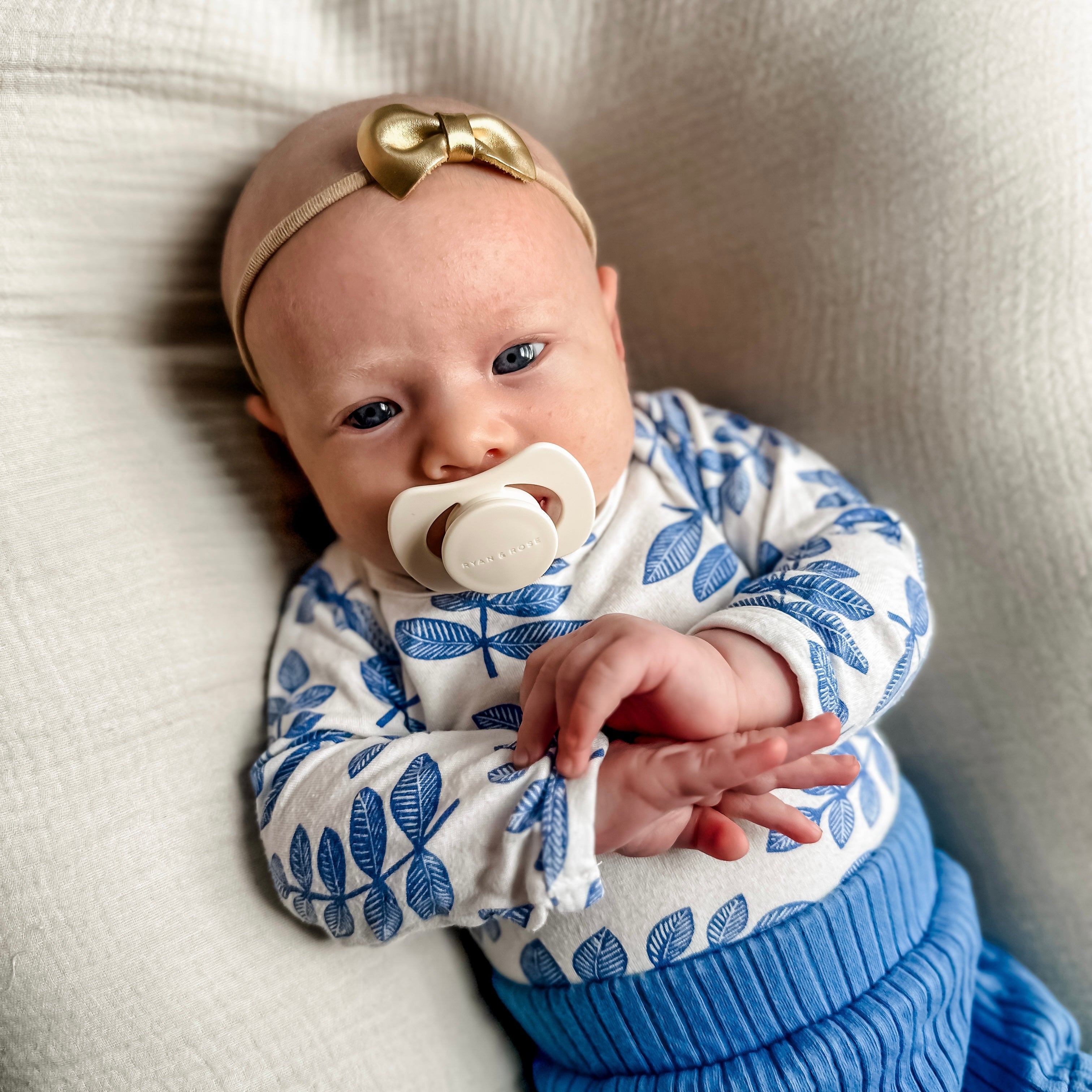 A baby wearing a blue and white patterned long-sleeve top, a blue ribbed knit bottom, a beige headband with a gold bow, and a pacifier, lying on a textured cream fabric.