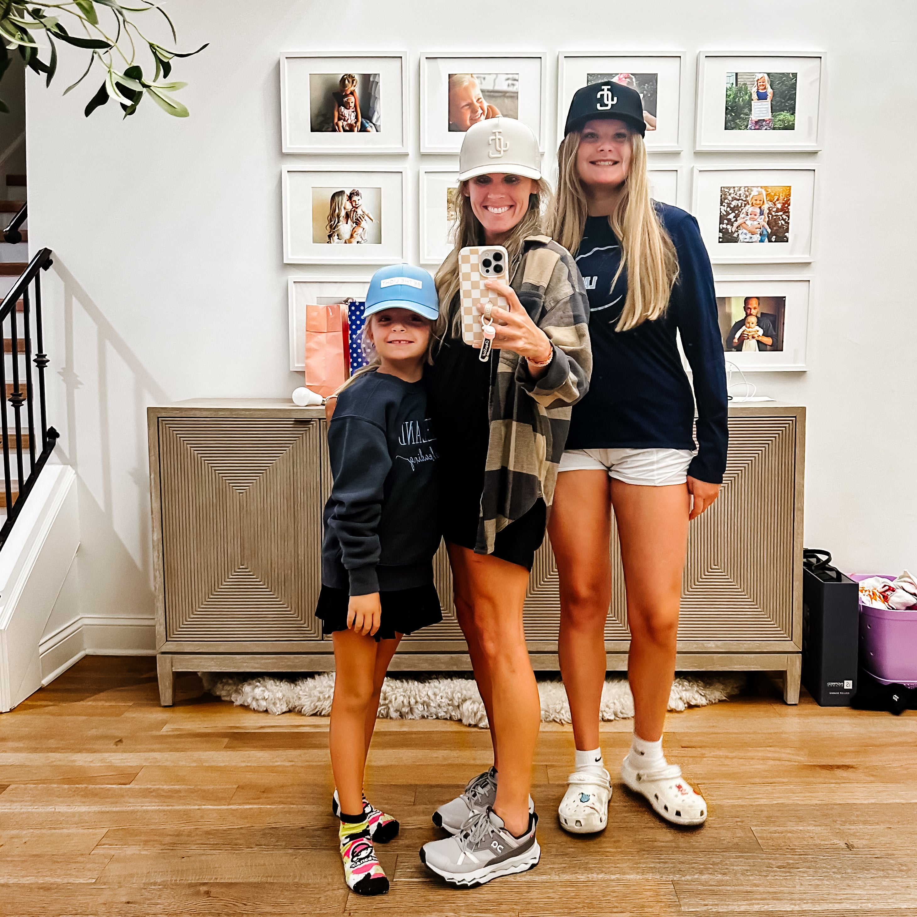 Three people taking a selfie in a home setting with framed photos on the wall.