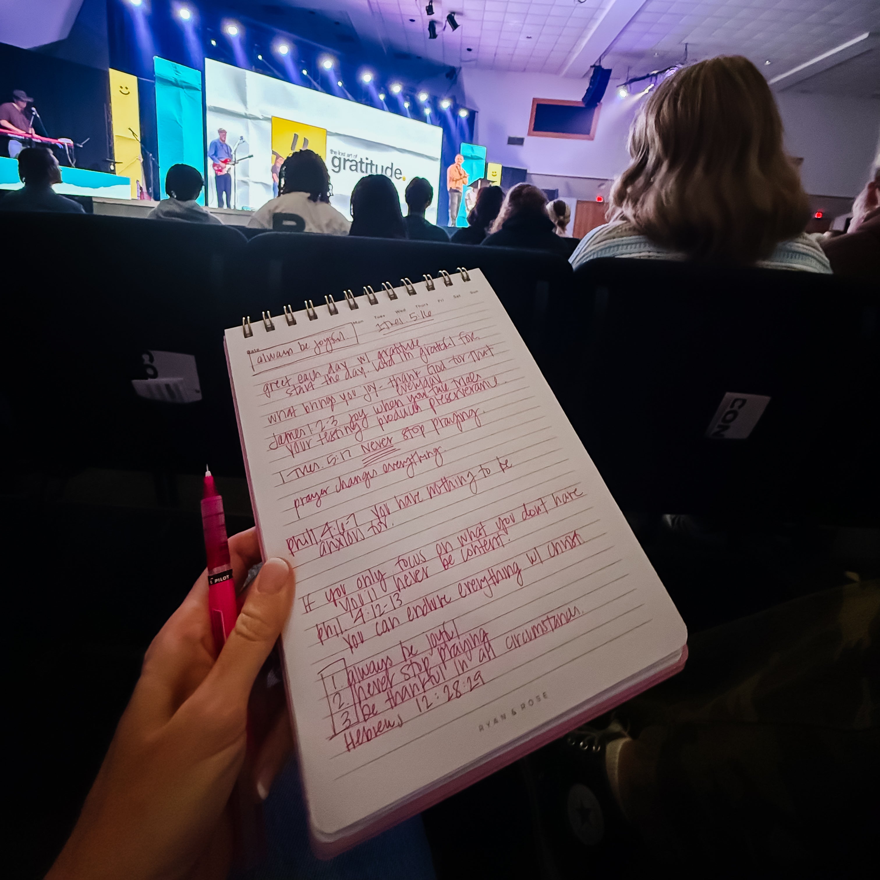 Person taking notes at a conference with a stage and audience in the background