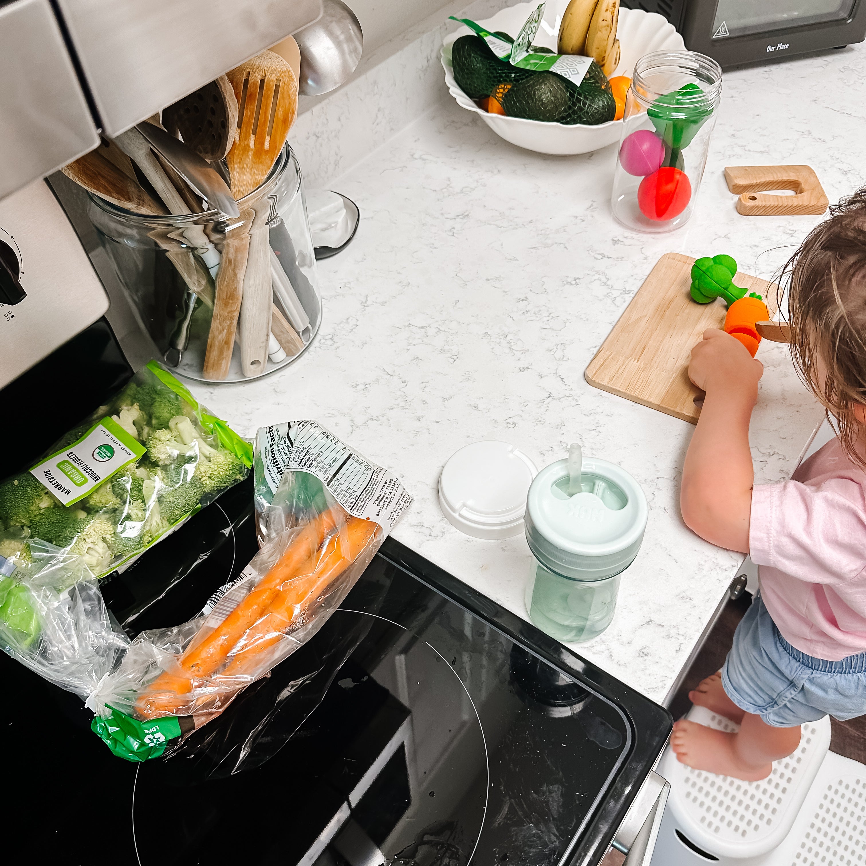 Child playing with toy vegetables on a kitchen counter next to real vegetables and kitchen appliances.