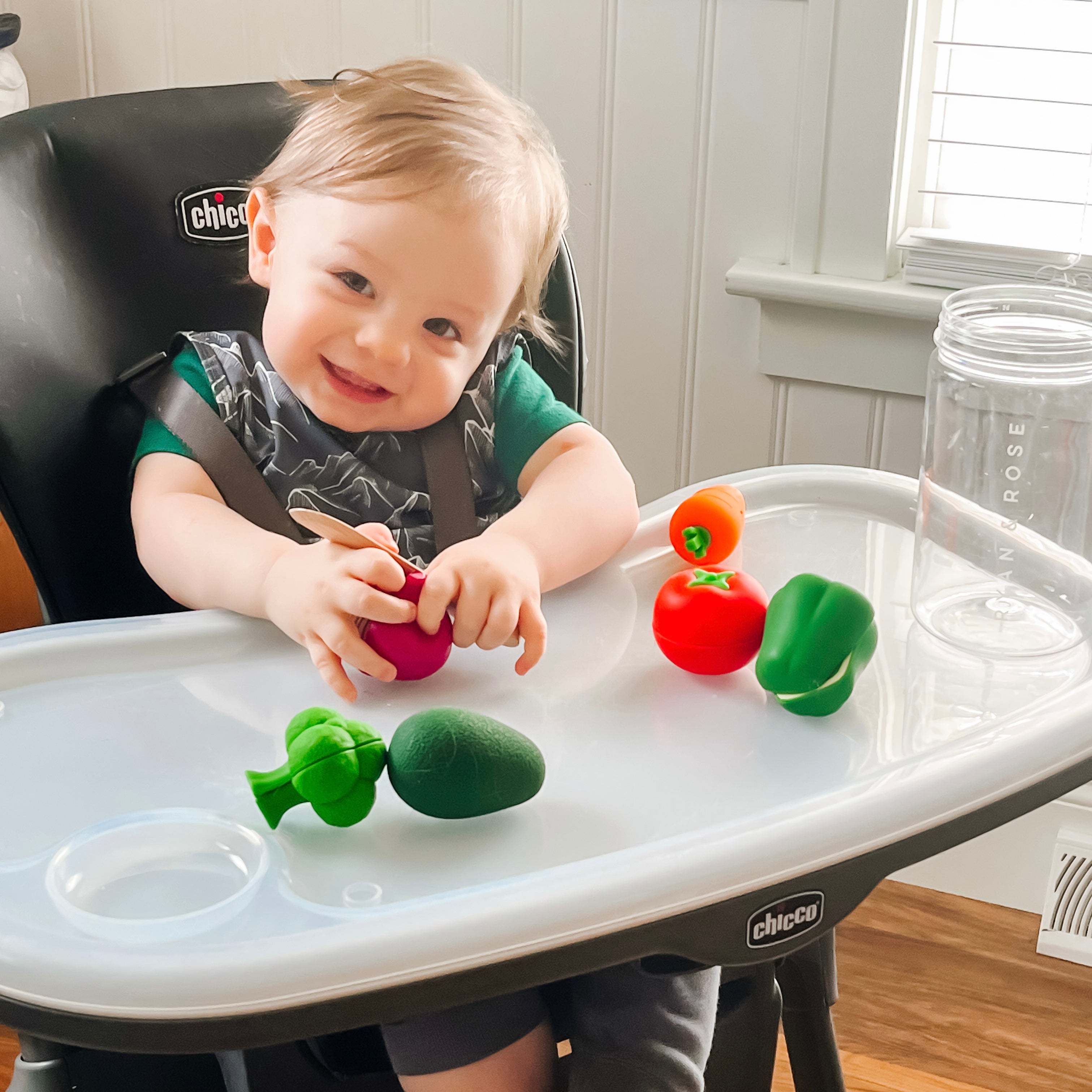 Child playing with colorful toy vegetables on a high chair tray.