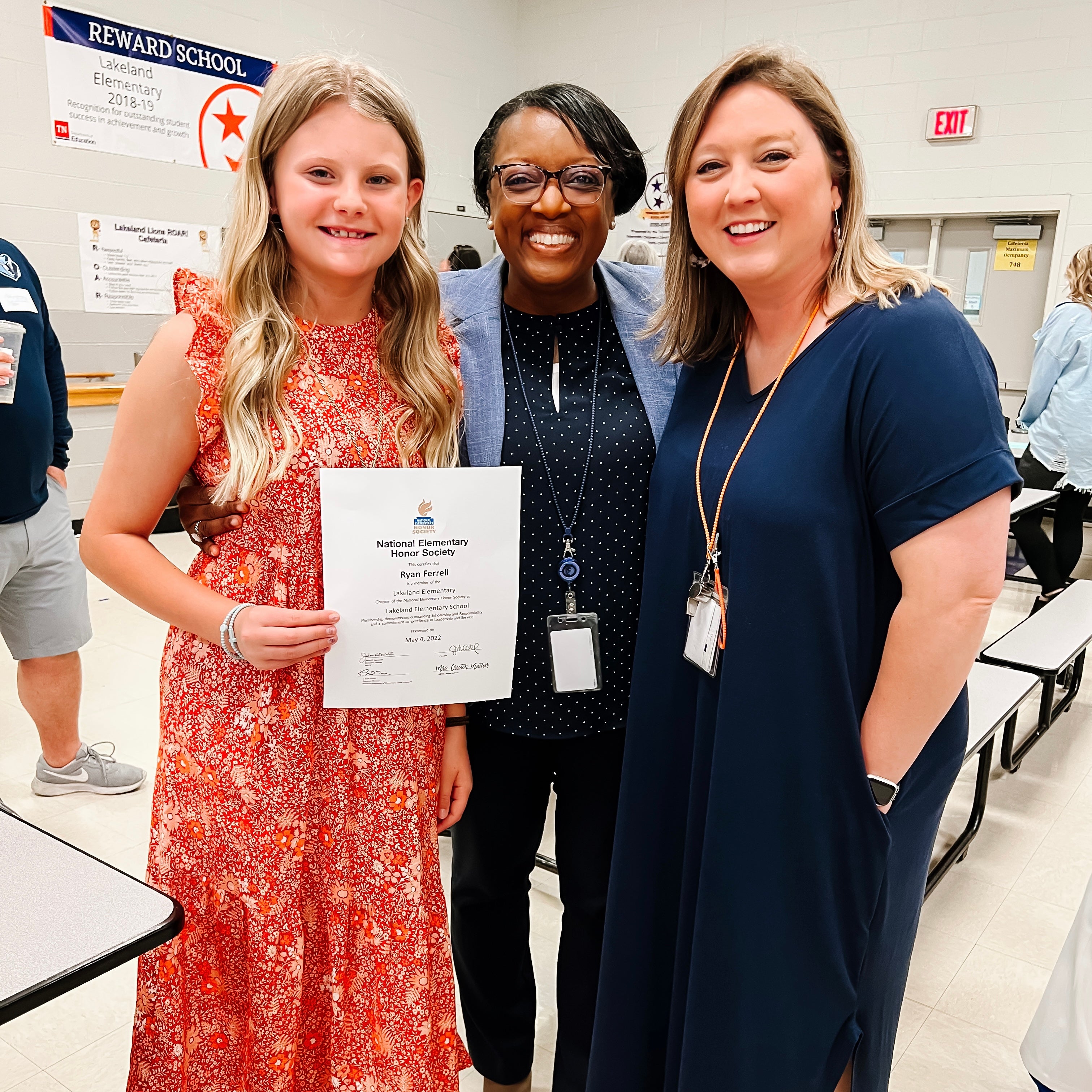 Girl in a red dress holding a certificate with two women in an indoor setting, possibly a school.