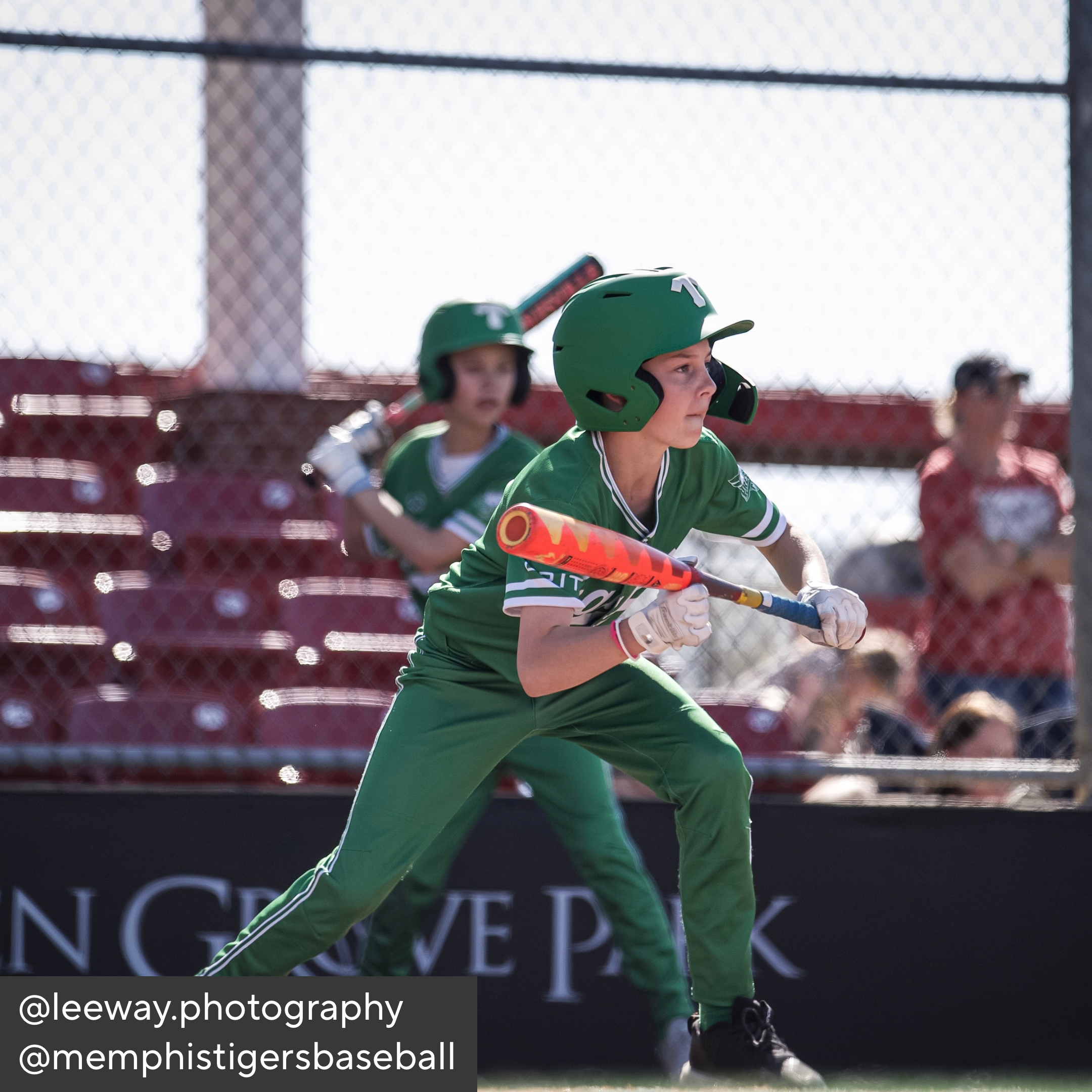 Child in green baseball uniform holding a bat on a field with spectators in the background.