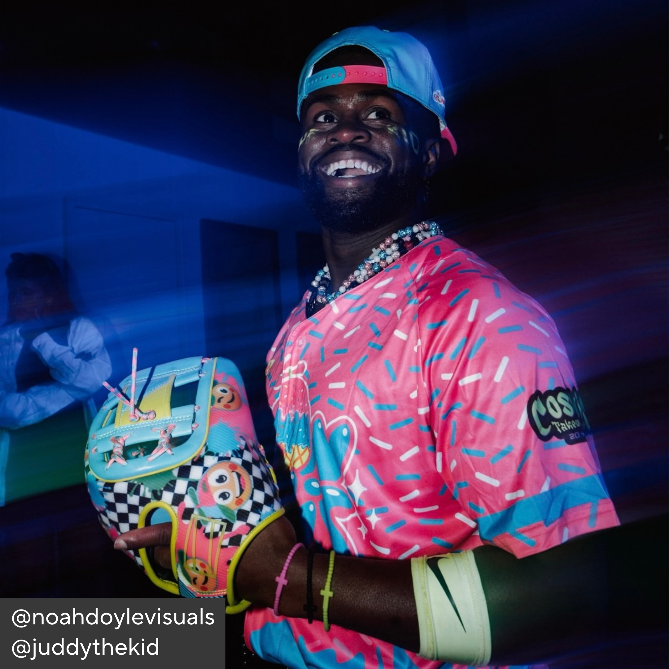 Person wearing a colorful outfit with a pink shirt and multicolored cap, holding a checkered helmet.