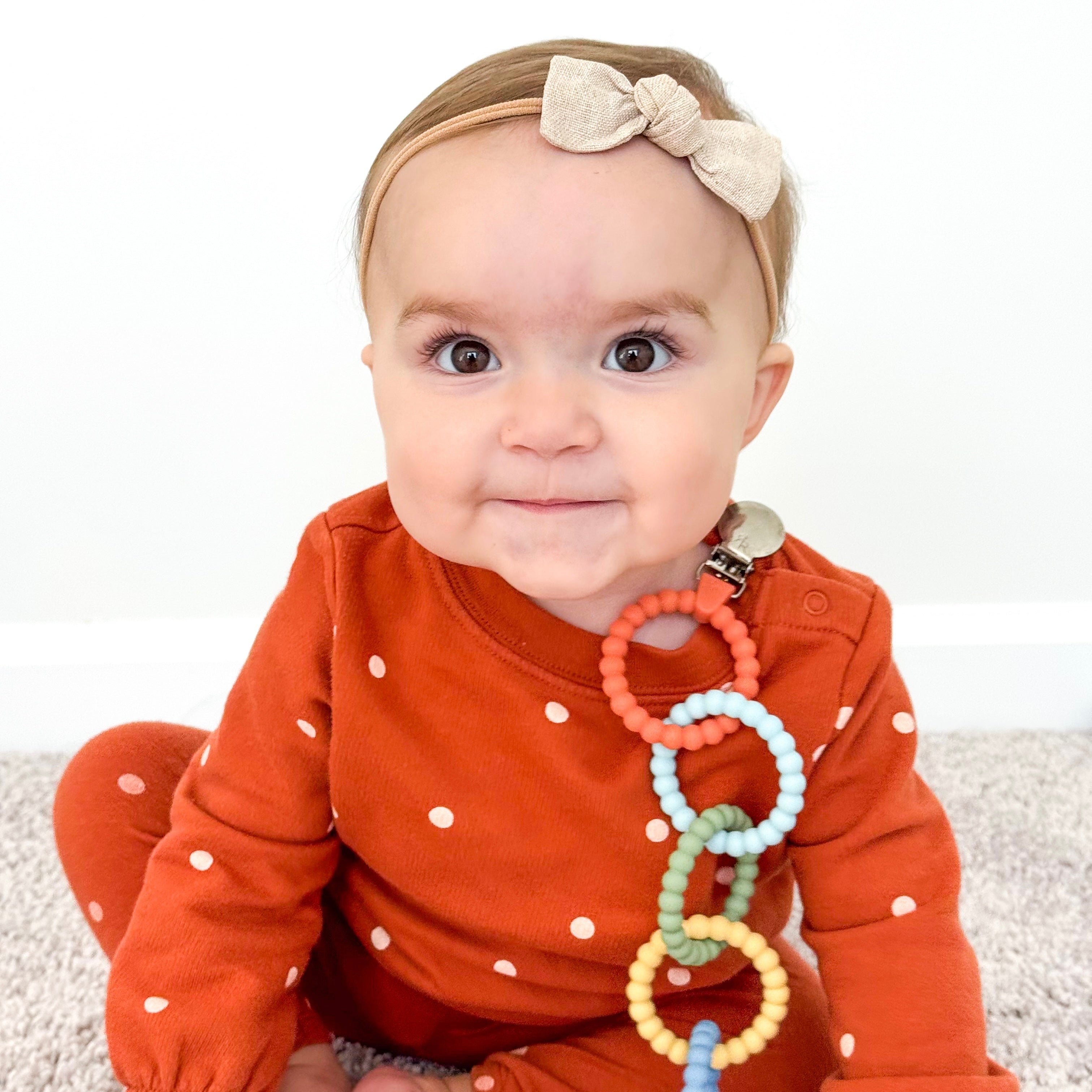 Baby in an orange outfit with a teething ring, sitting on a white surface.