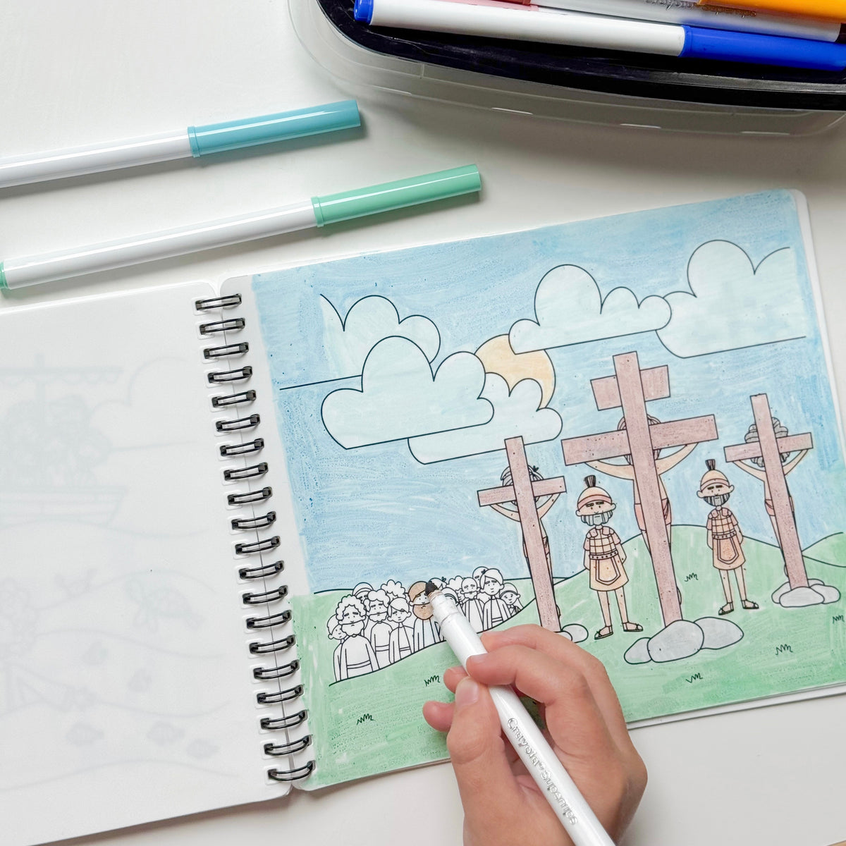 A hand holds a white marker over an open spiral notebook showing a colorful drawing of crosses, people, and clouds. Two markers lie on the white surface above the notebook, with a pen case partially visible at the top.
