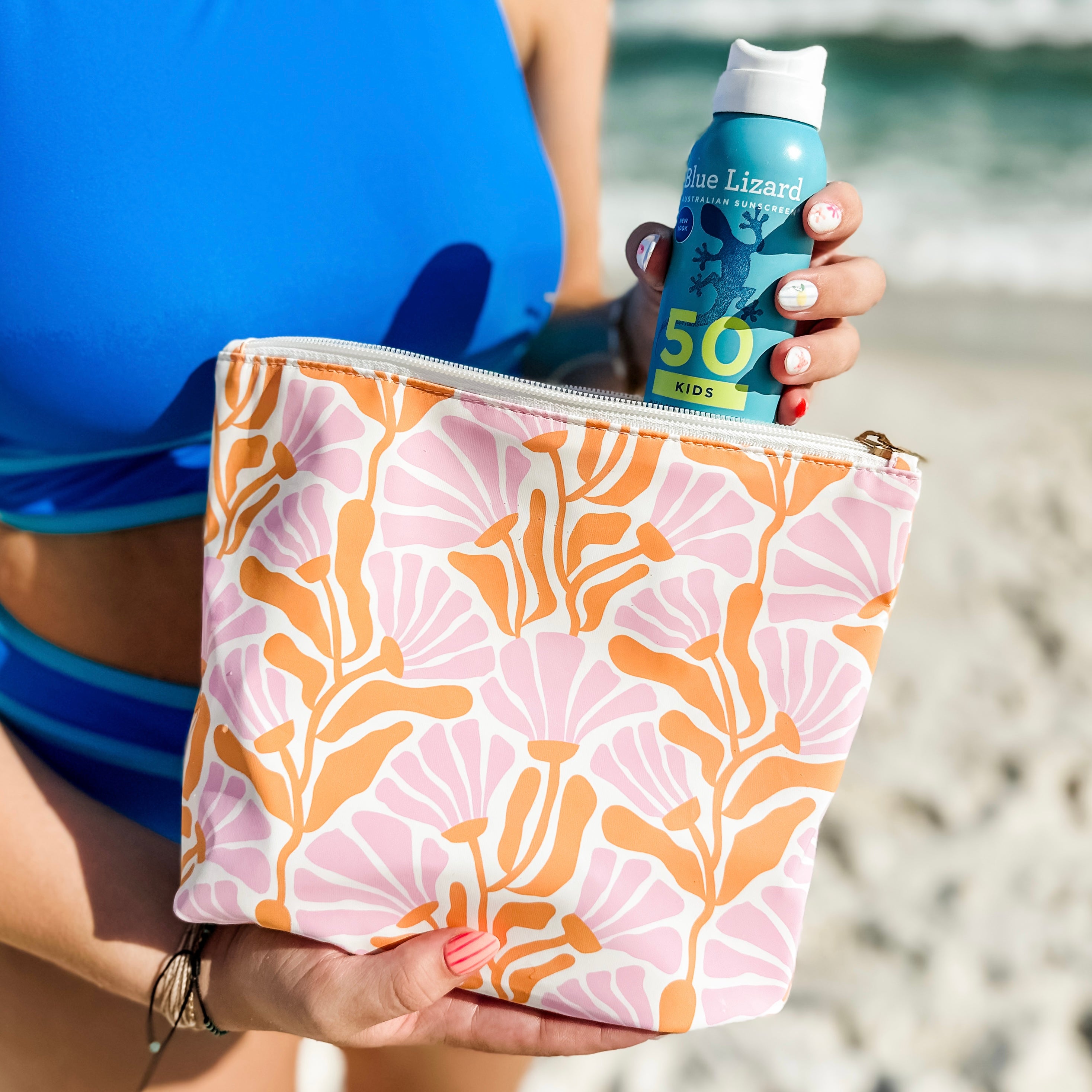 Person holding a Blue Lizard sunscreen bottle and a patterned pouch on a beach.