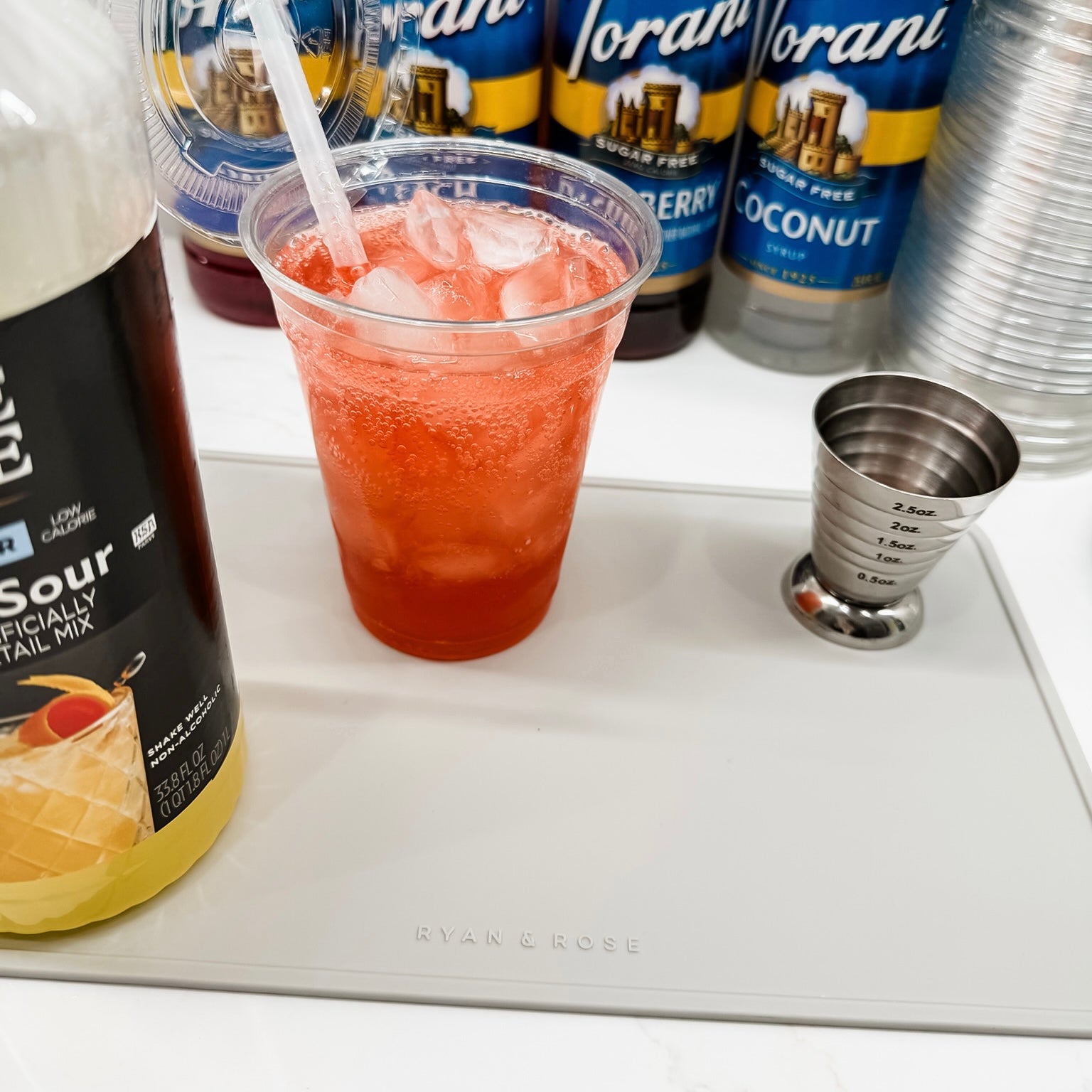 Red cocktail in a clear cup with ice, surrounded by Torani syrup bottles and a bottle of Bitter Sours mix.