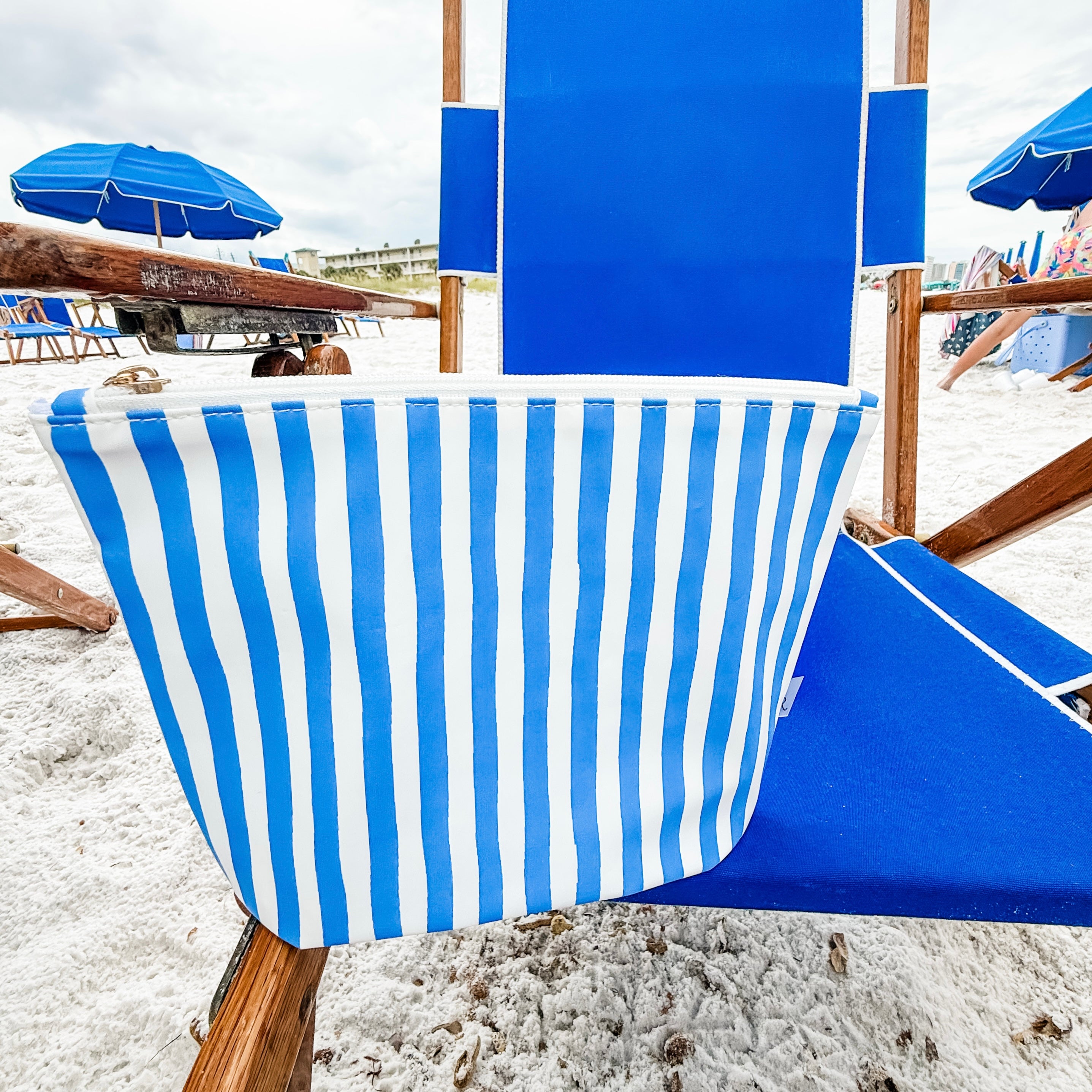 Blue and white striped beach chair on a sandy beach with umbrellas in the background.