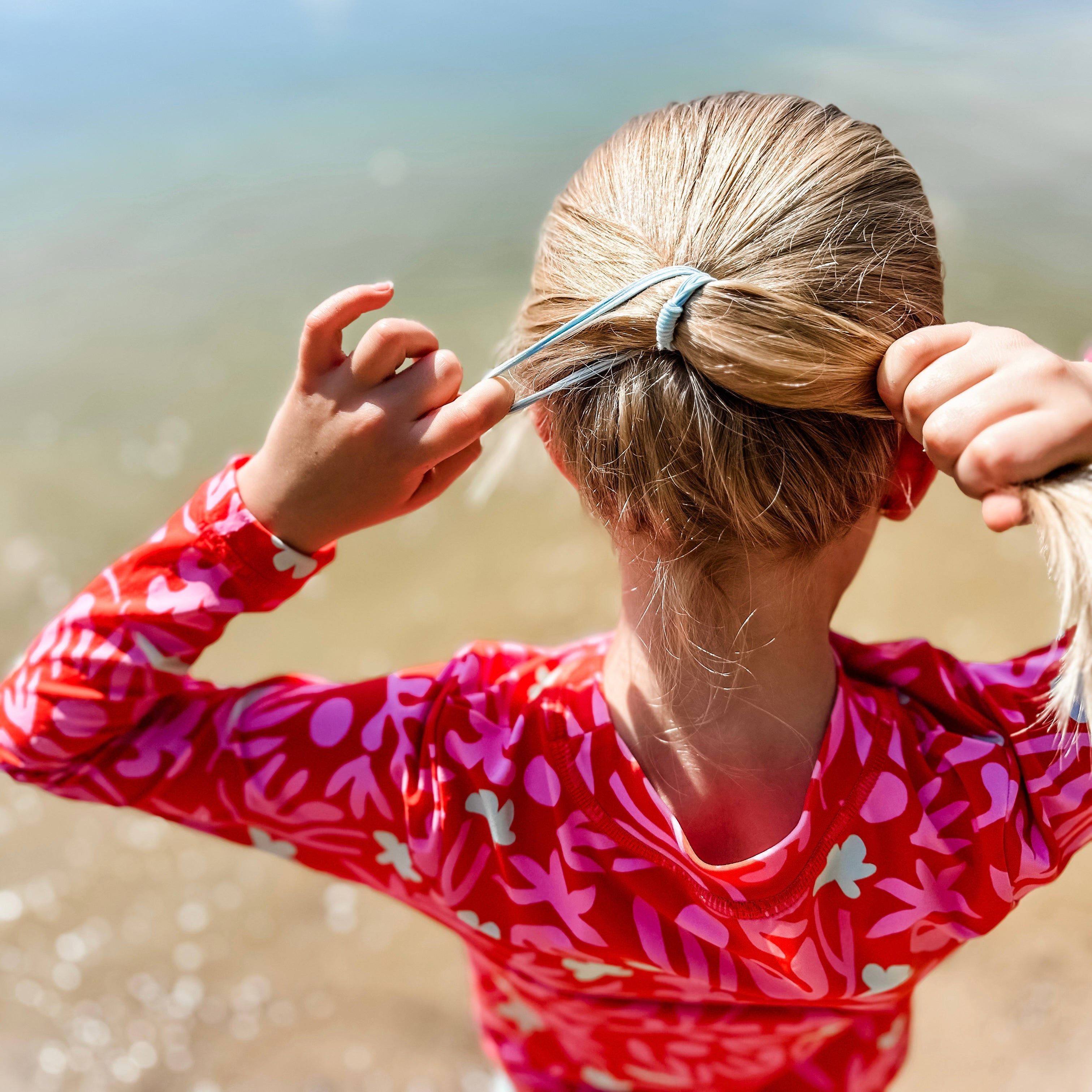 Person in a red and pink patterned shirt tying their hair on a beach.