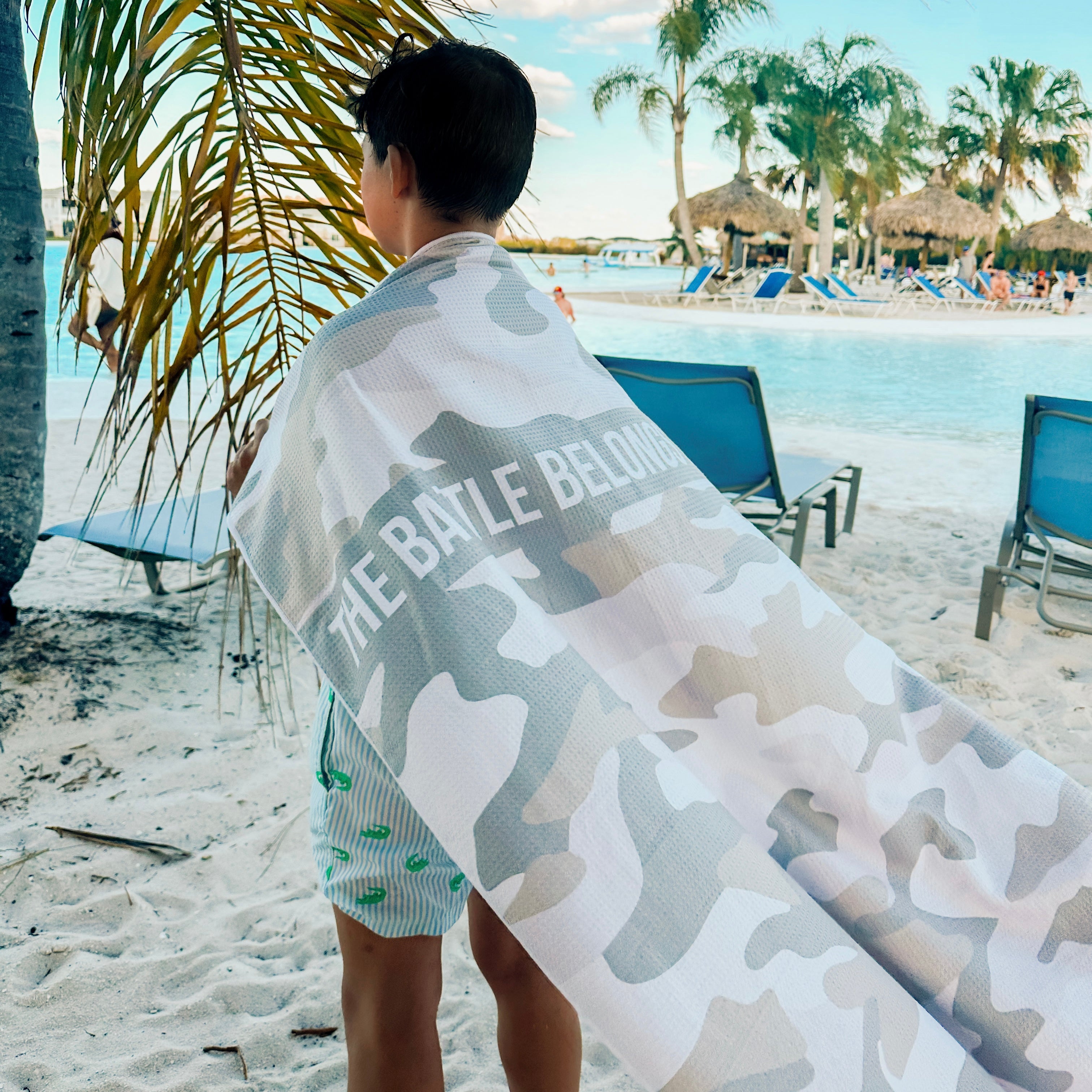 Person on a beach with a camouflage towel draped over them, featuring the text &