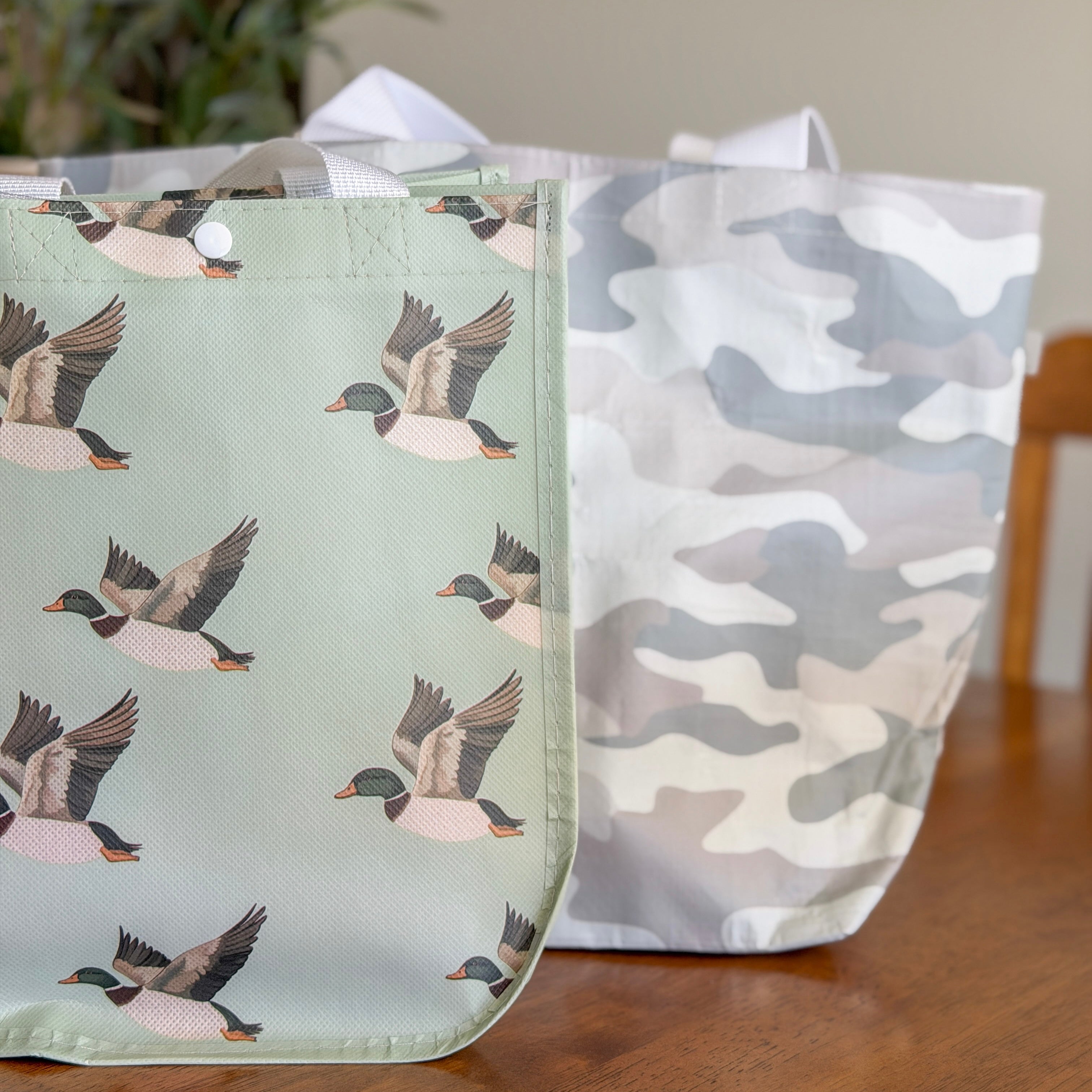 Two reusable shopping bags, one with duck pattern and the other with camouflage design, on a wooden surface.