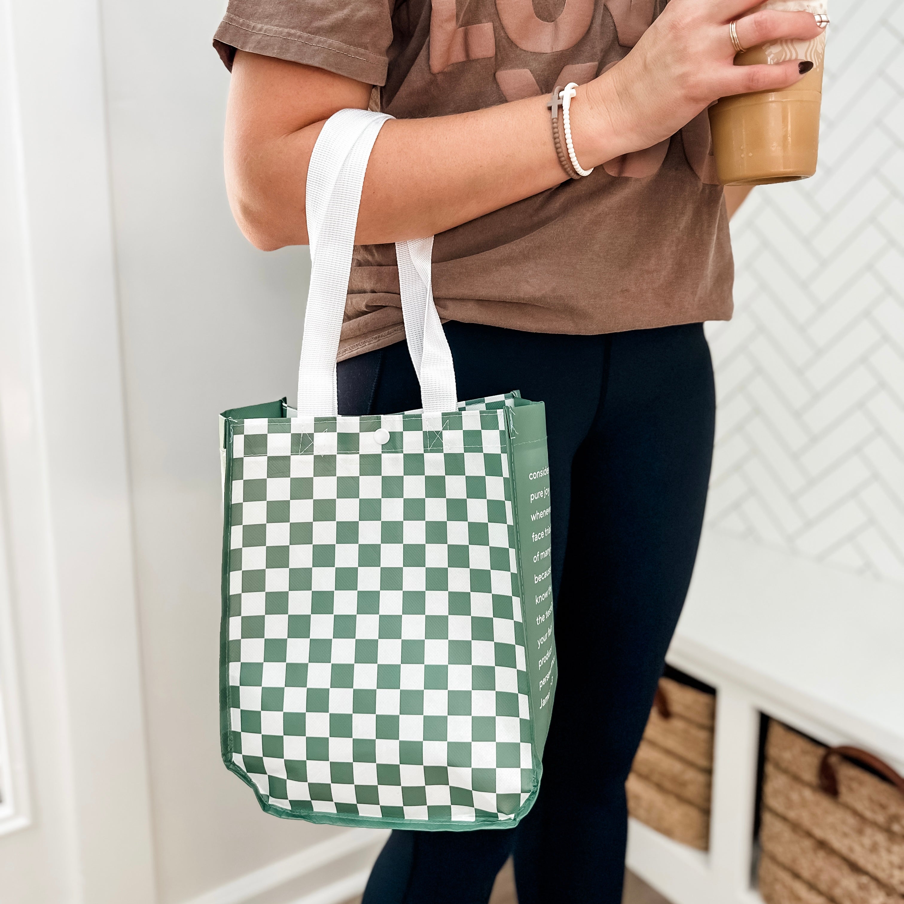 Person holding a green and white checkered tote bag and a coffee cup indoors.