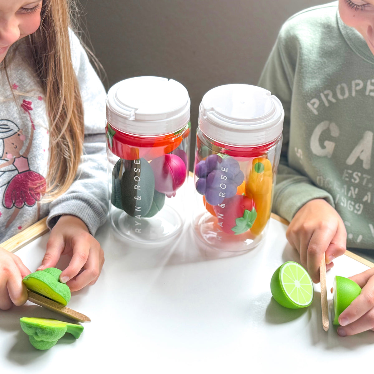Two children cutting green fruit props inside clear plastic cups with white lids, set on a white tabletop.