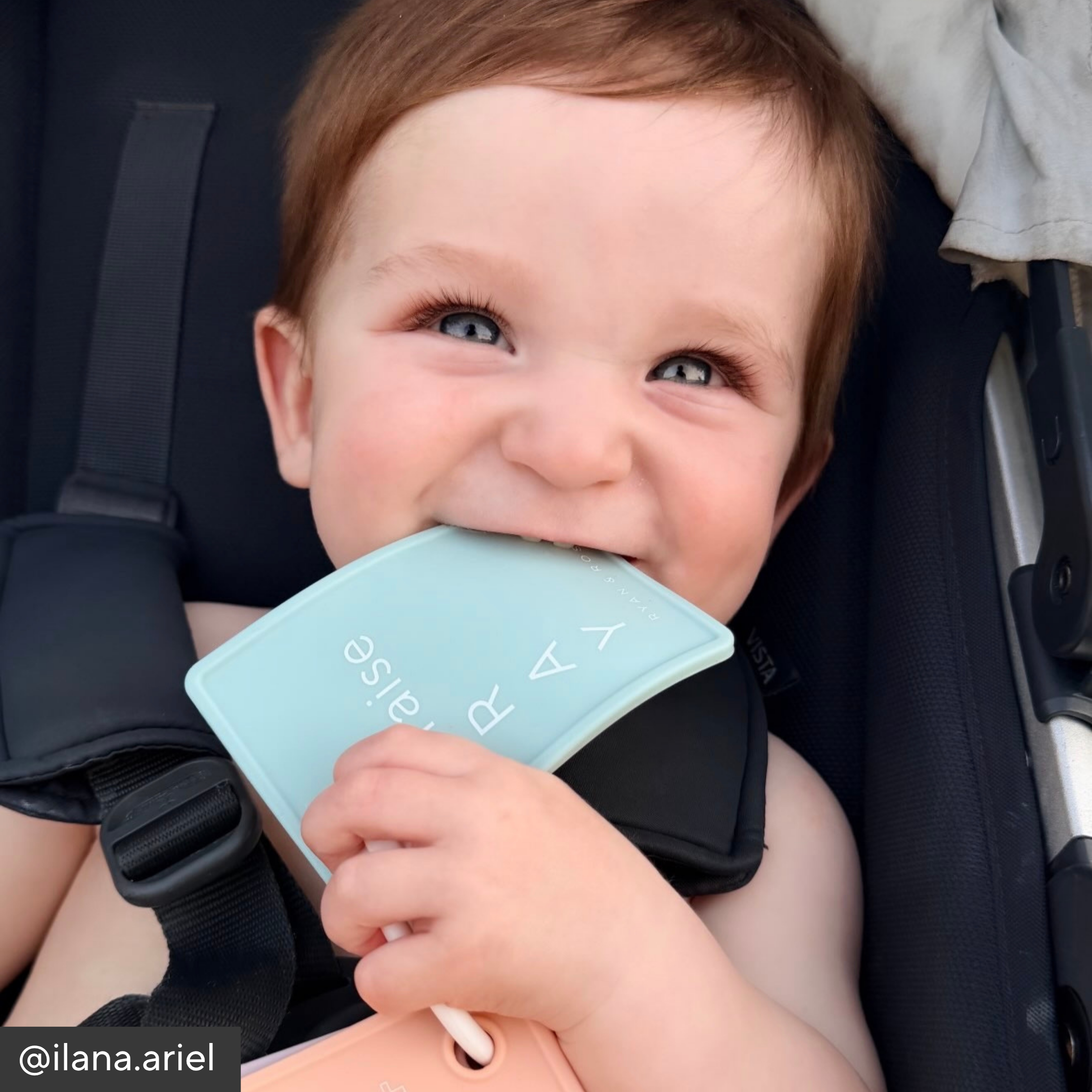 A baby wearing a black stroller seat belt and holding a light blue silicone shield with the brand name &
