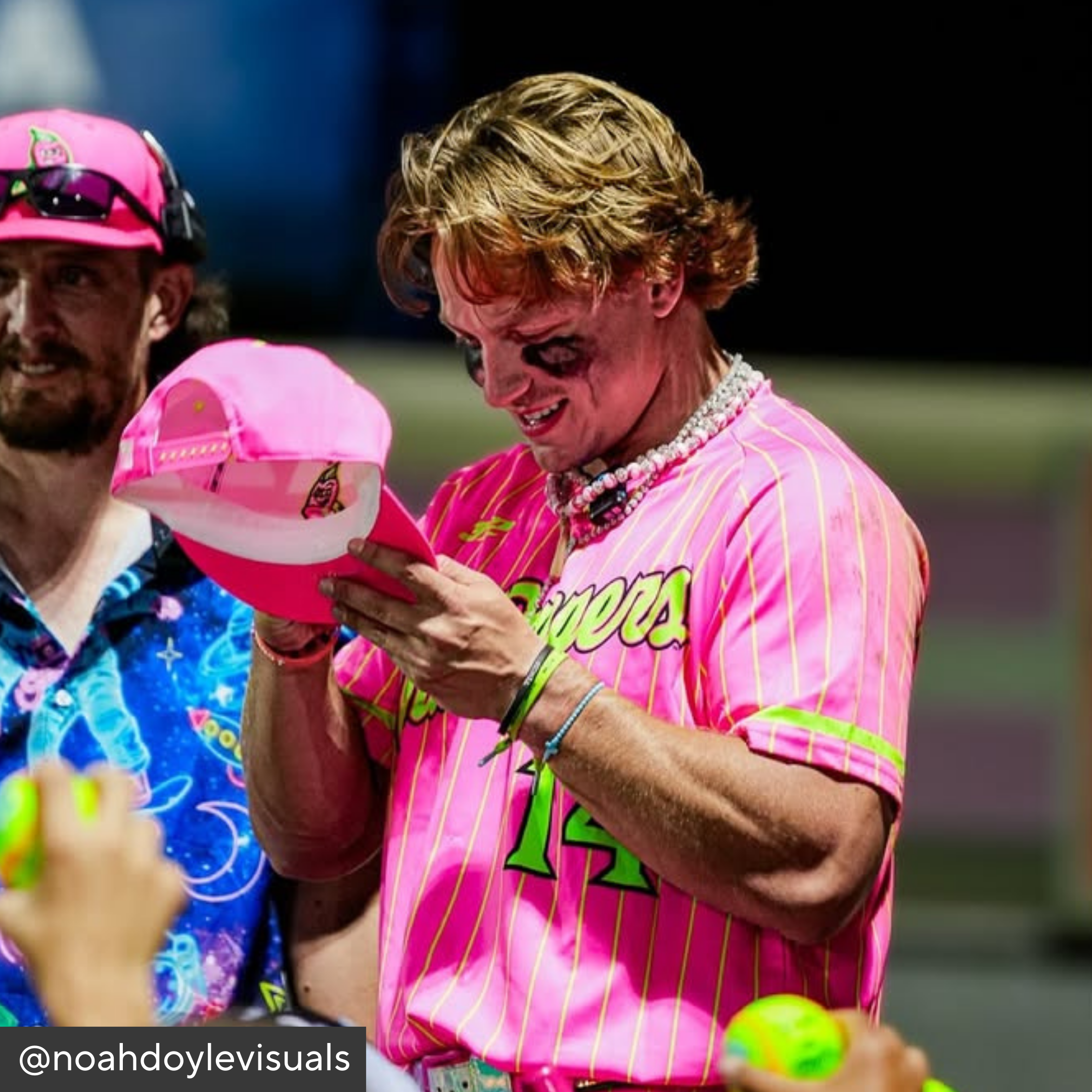 Person in a pink baseball uniform holding a pink cap with a cartoon character, surrounded by others.