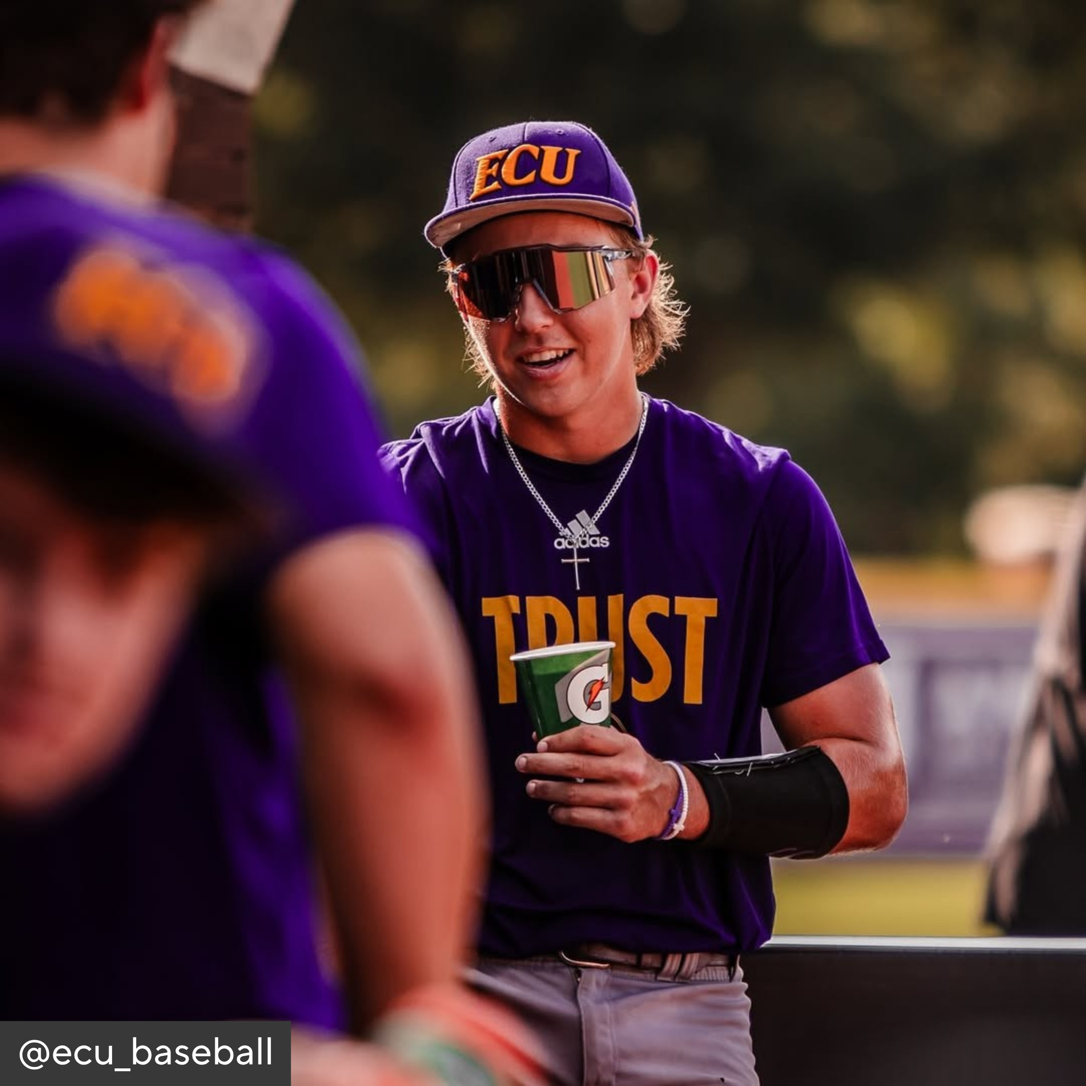 Person wearing an ECU cap and sunglasses, holding a green cup with a logo, surrounded by others in purple shirts.