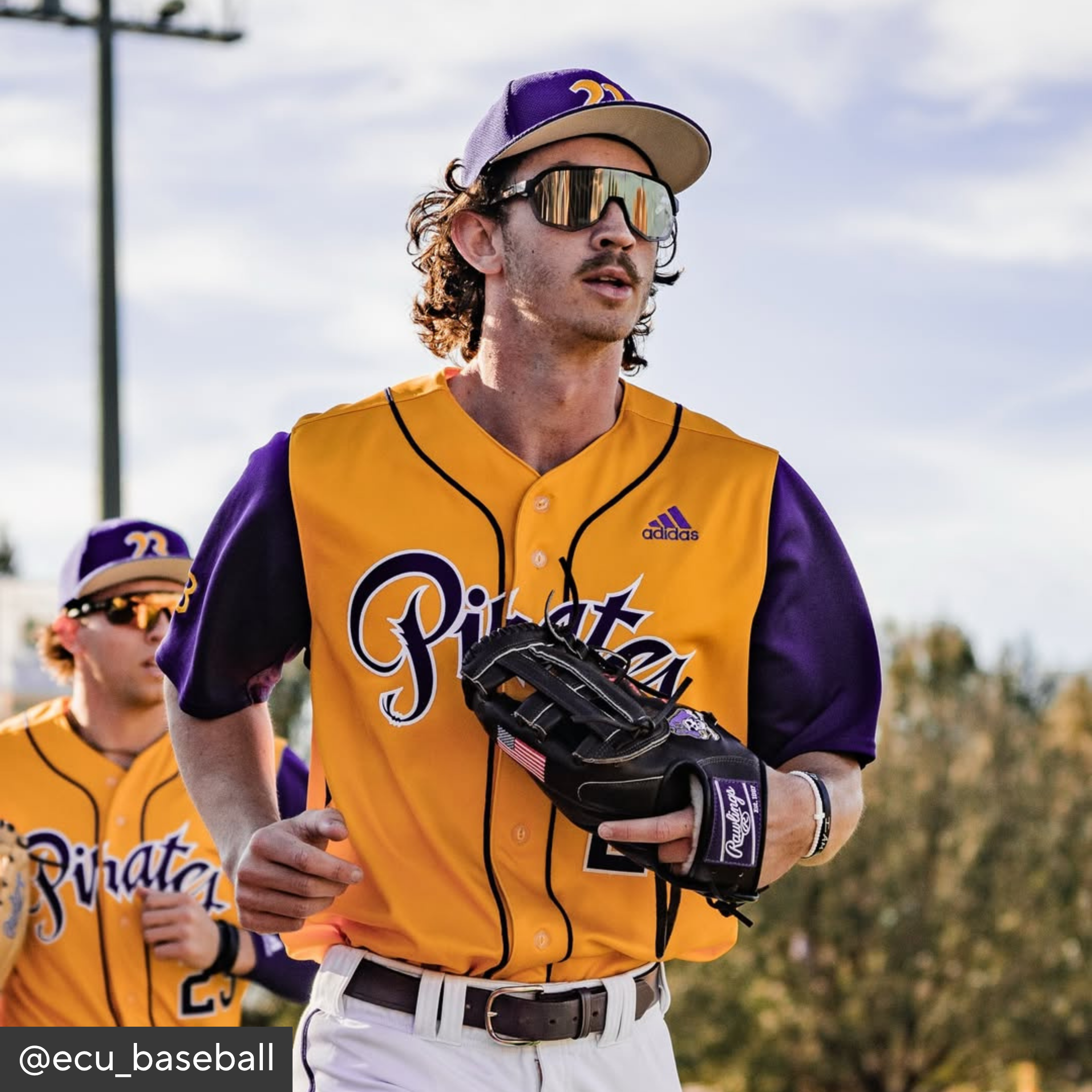 Baseball player in yellow and purple uniform with sunglasses and glove, standing outdoors.