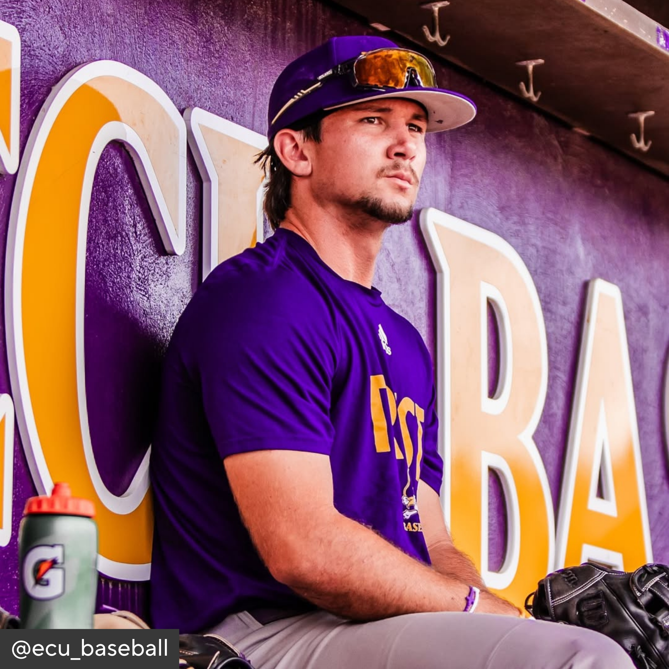 Baseball player in a purple uniform sitting in a dugout with a Gatorade bottle and glove.