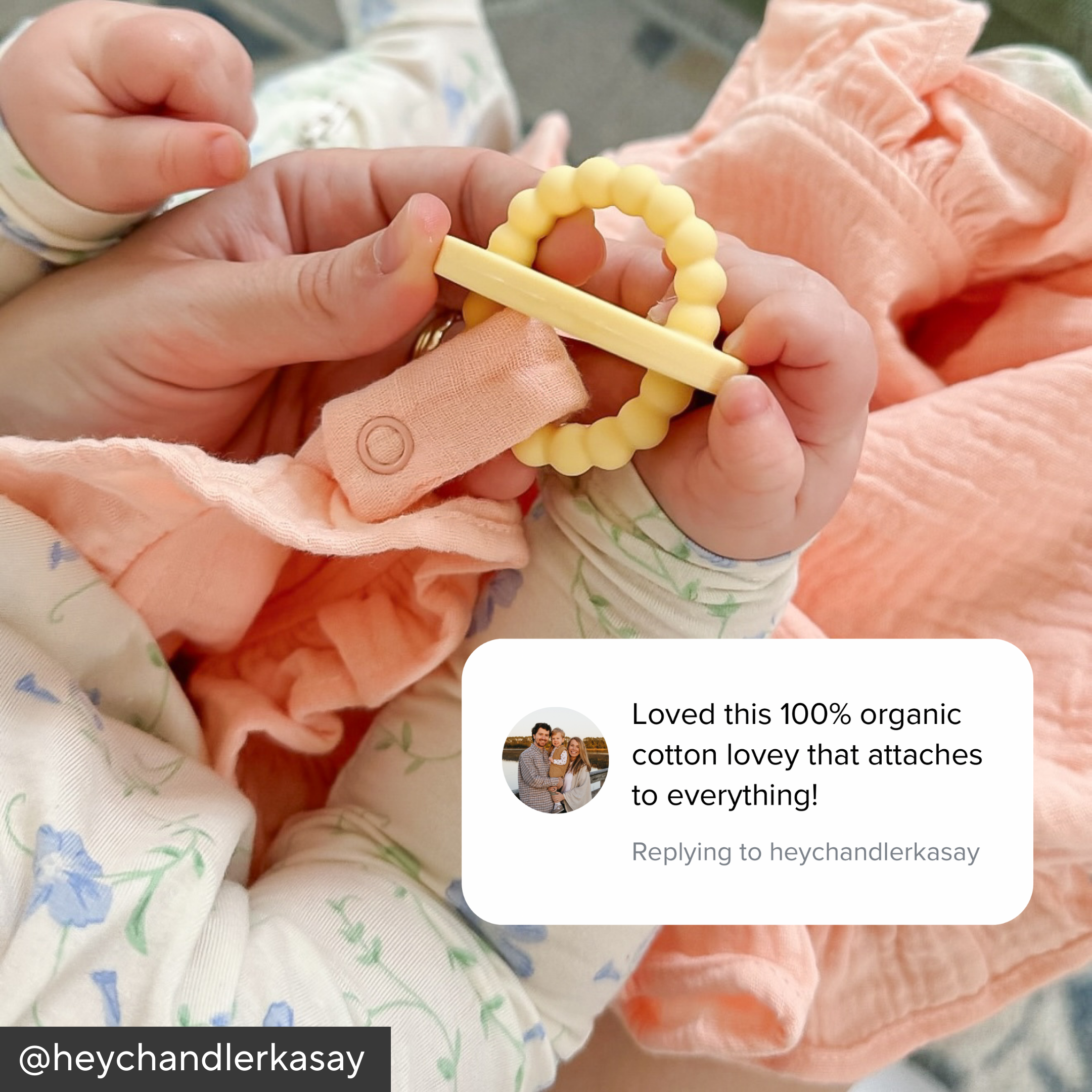 Baby holding a yellow teething ring with a pink blanket and floral-patterned fabric in the background.