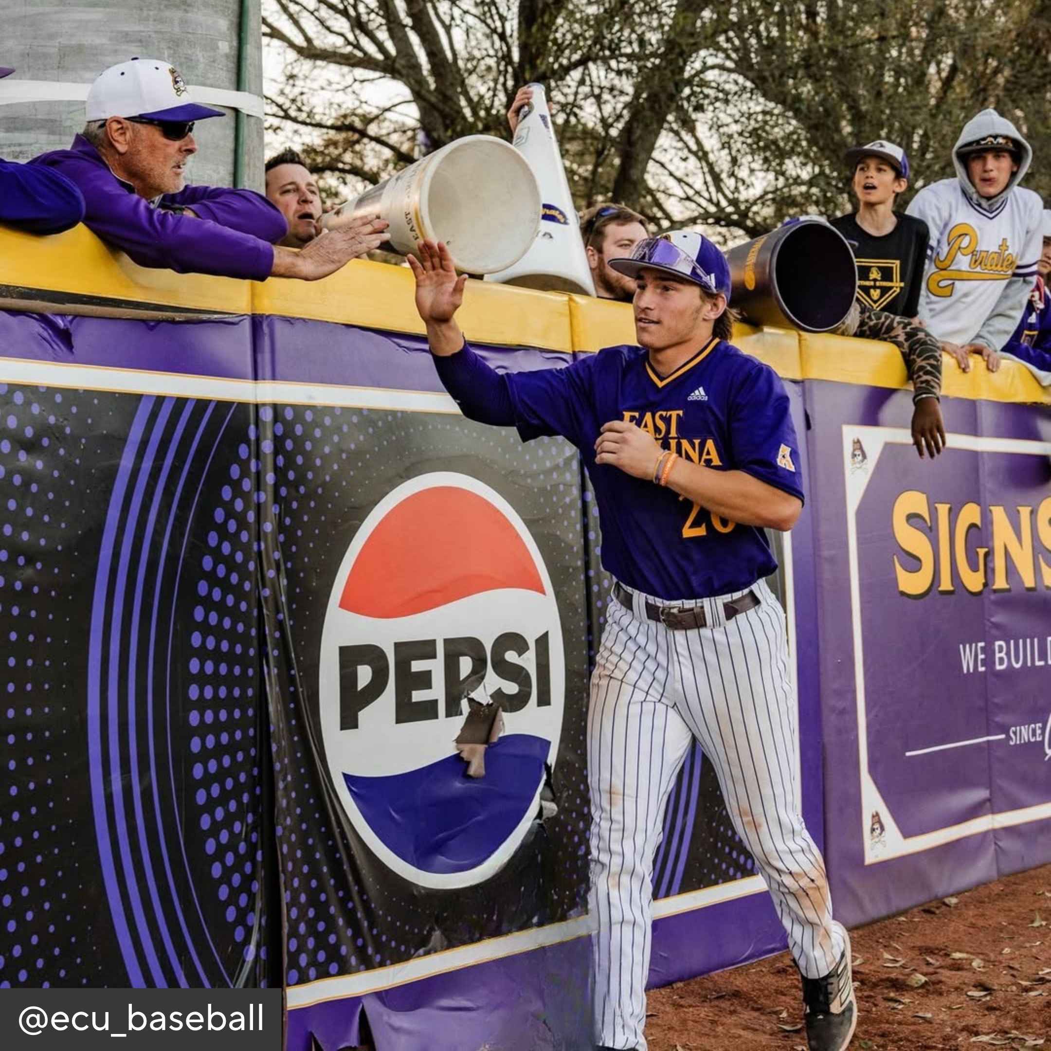 Baseball player in East Carolina uniform shaking hands with a fan in front of a Pepsi advertisement.
