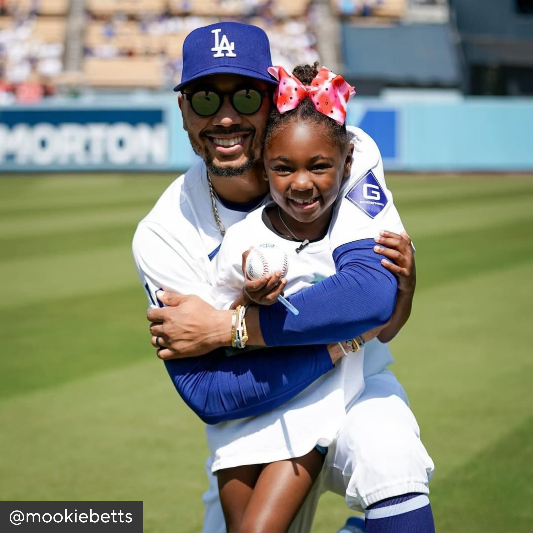 Man and young girl in baseball uniforms on a field, with the man holding a baseball.