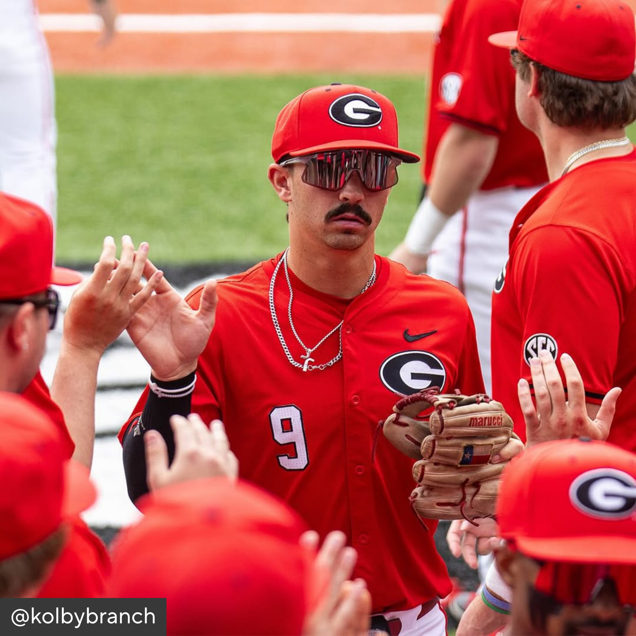 Baseball player in red uniform with sunglasses and a mustache interacting with teammates on a field.