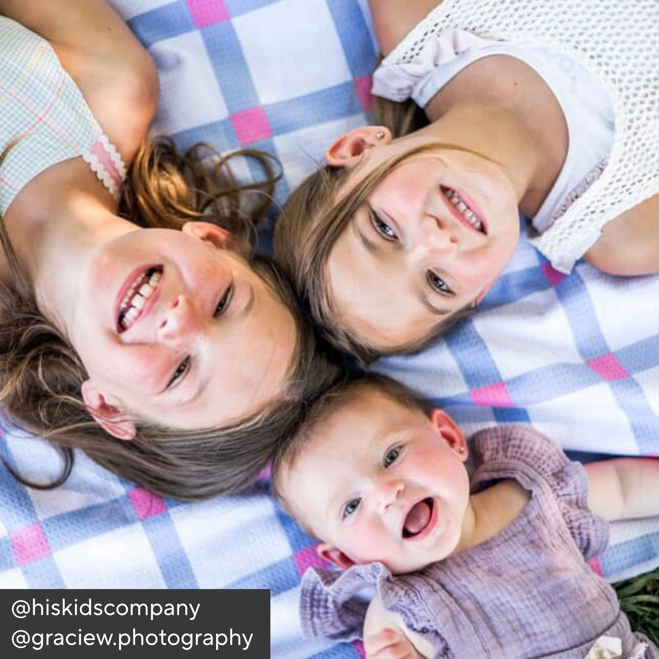 Three children lying on a checkered blanket, smiling upwards.