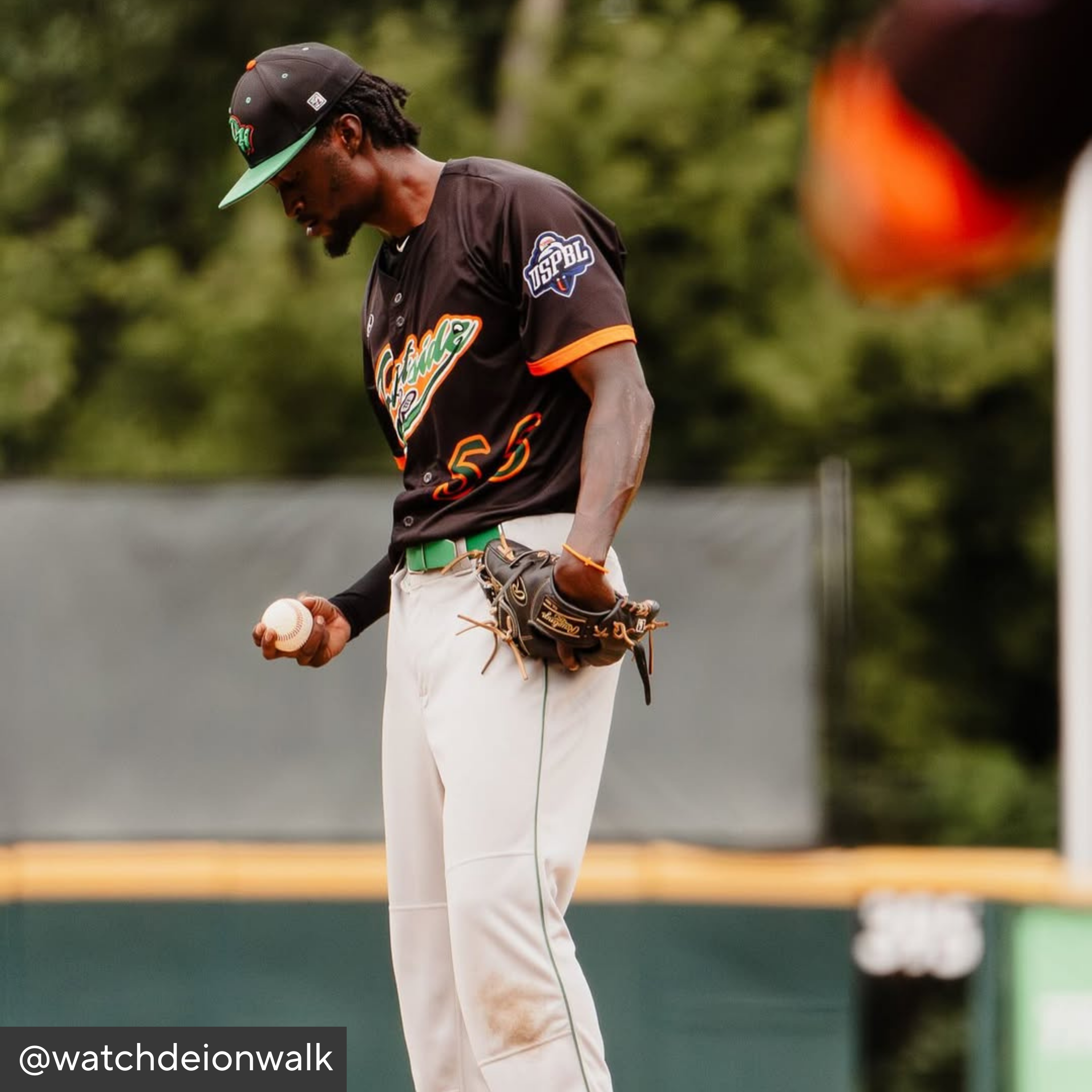 Baseball player in a black and orange uniform with a green cap, holding a baseball and glove on a field.