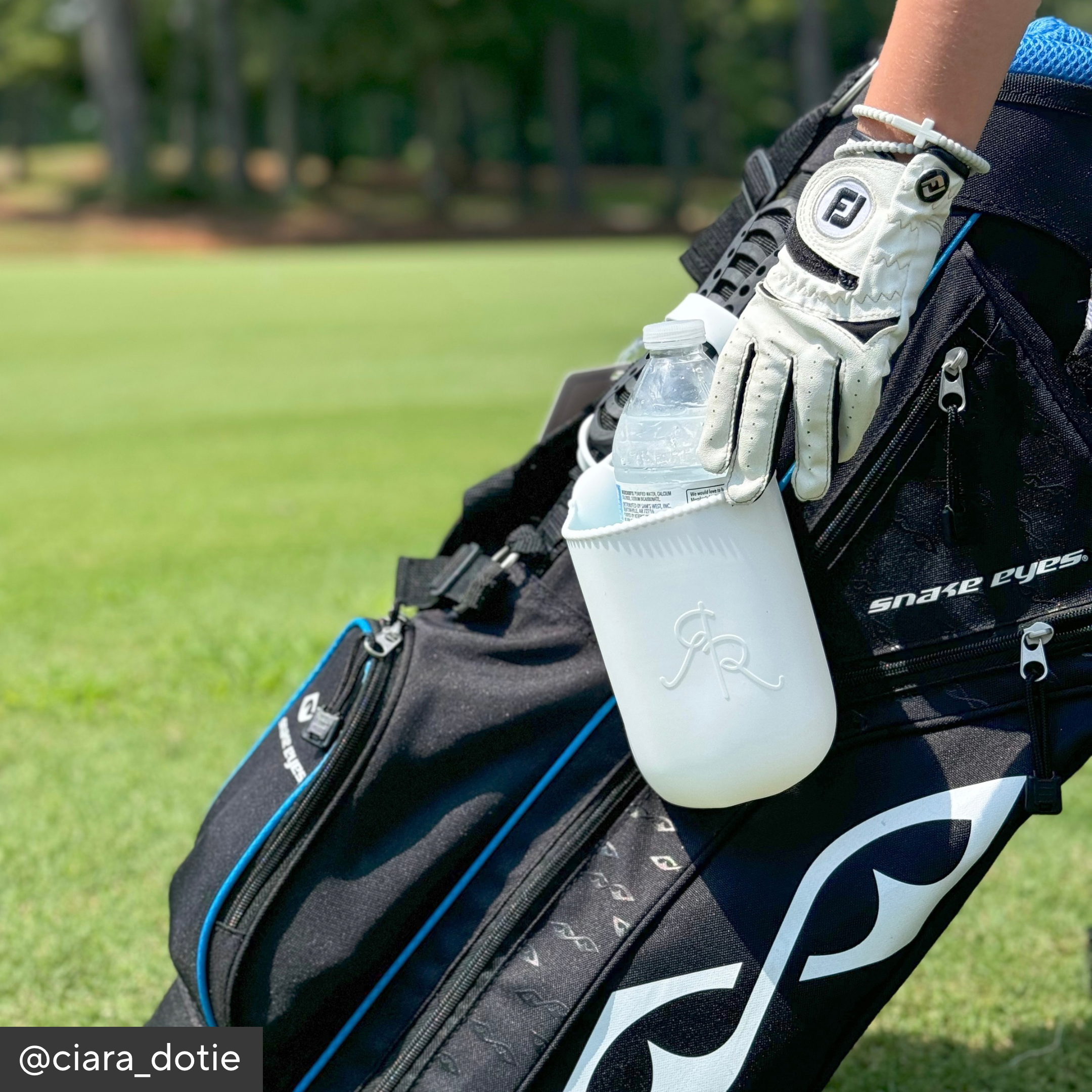 Person holding a water bottle with a golf bag and club in the background on a golf course.