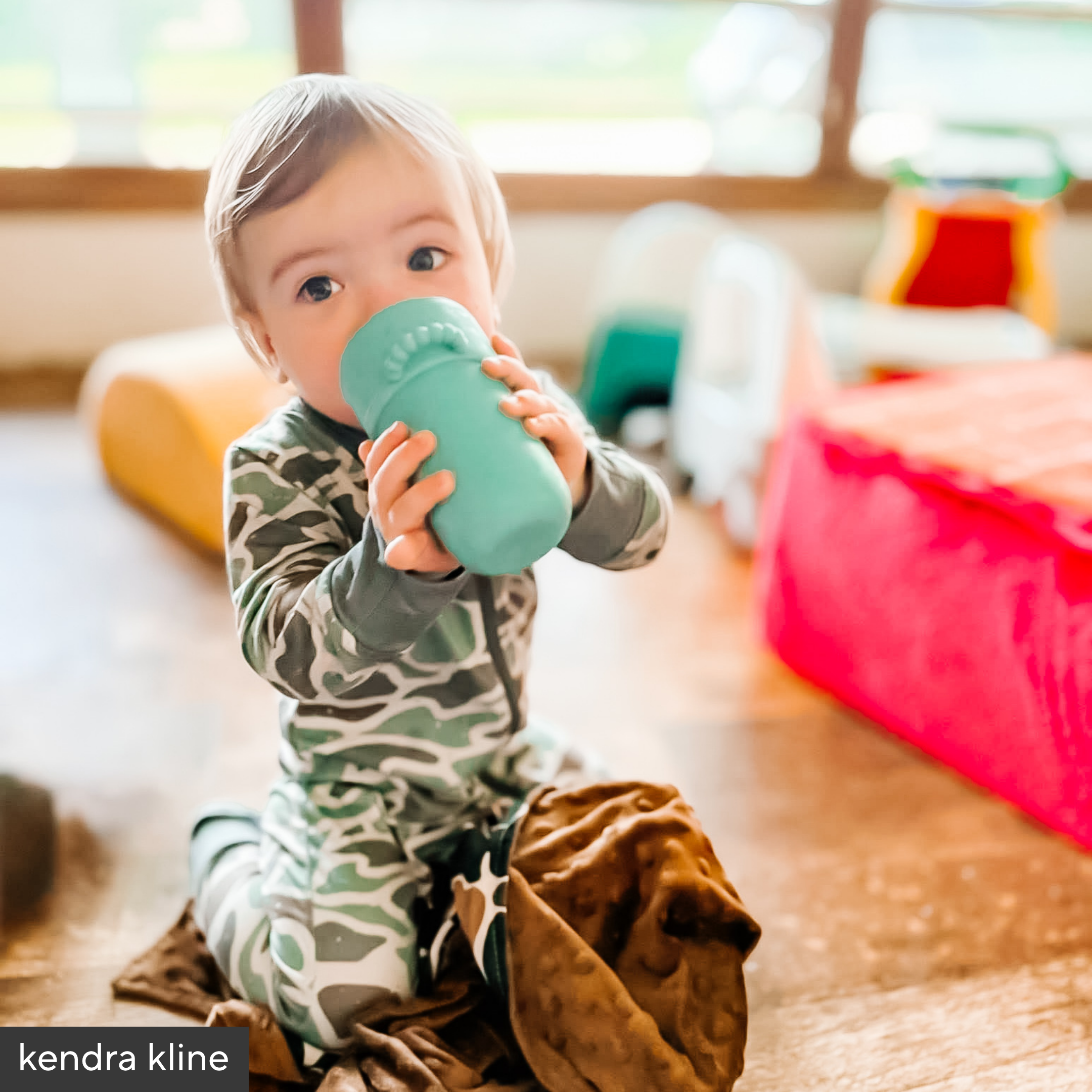 Child holding a green sippy cup in a room with colorful toys