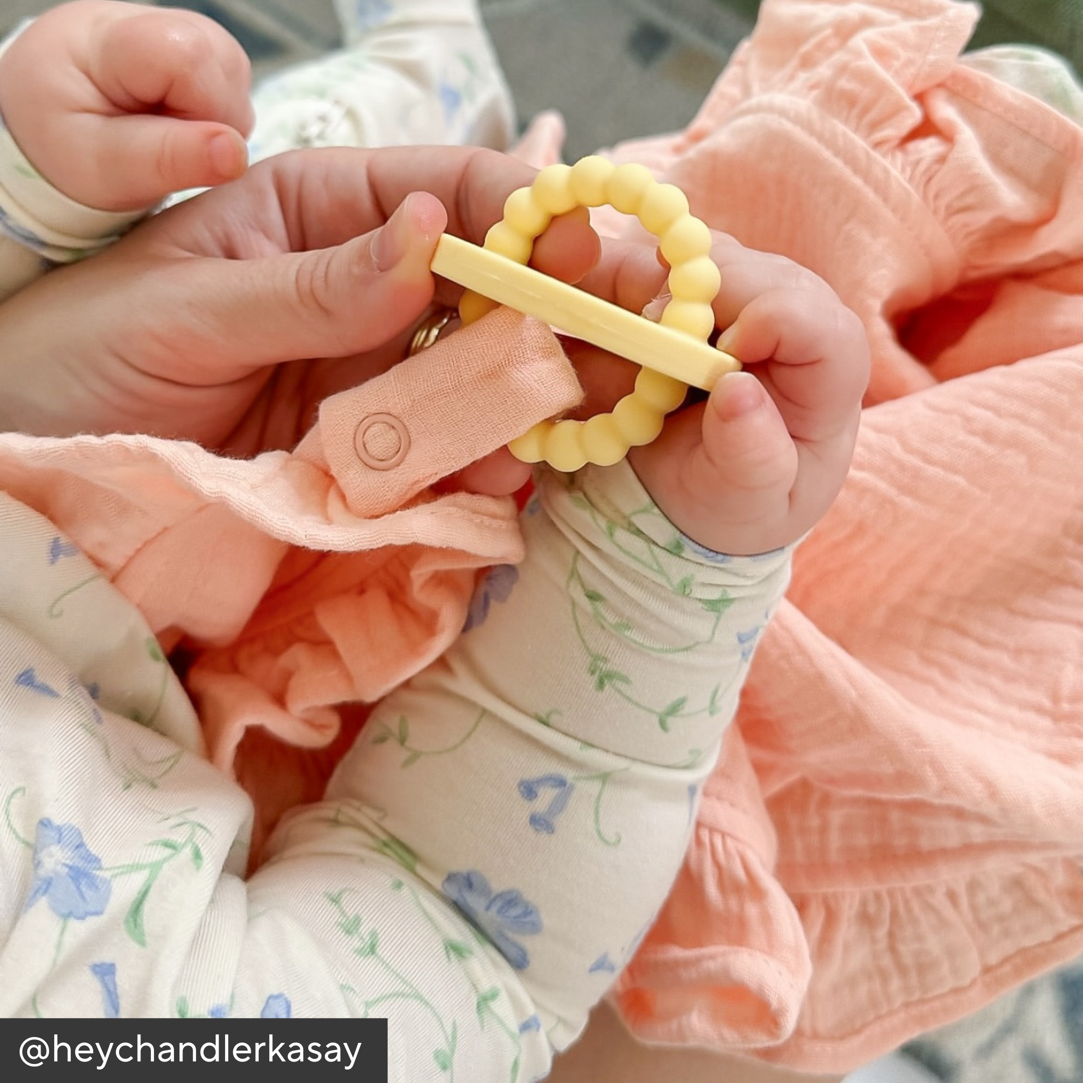 Baby holding a yellow teething ring with a pink blanket and floral-patterned fabric in the background.