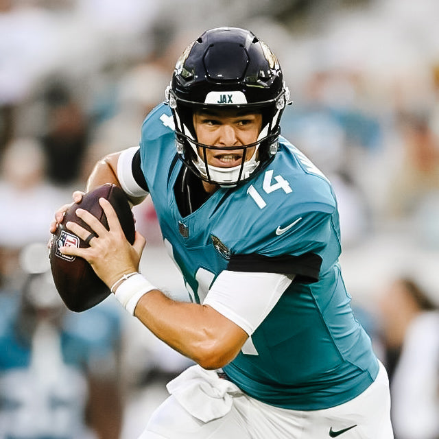 Football player in teal jersey with number 14 holding a ball, blurred crowd in the background