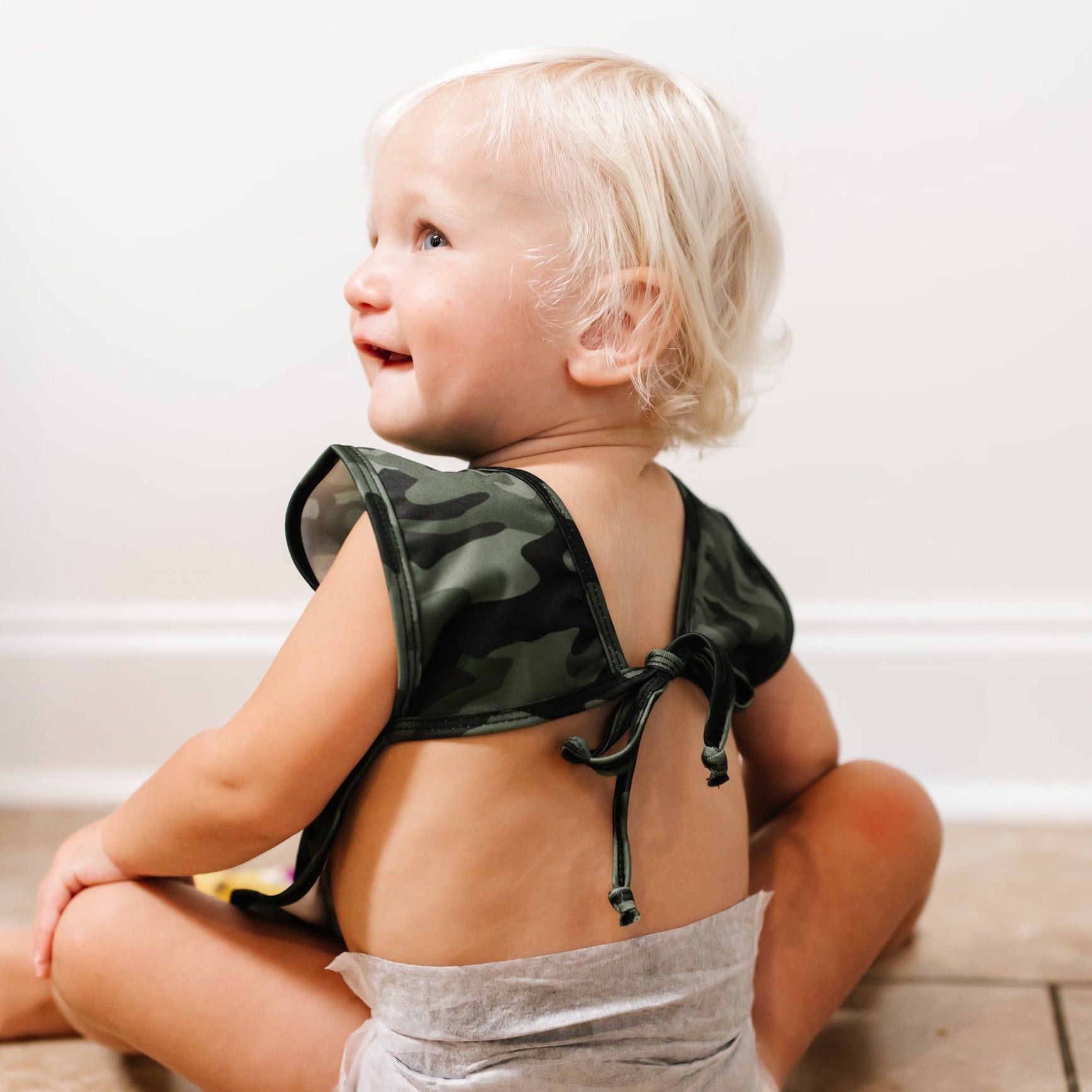 Baby sitting on the floor wearing the Camo Cutie Bapron.