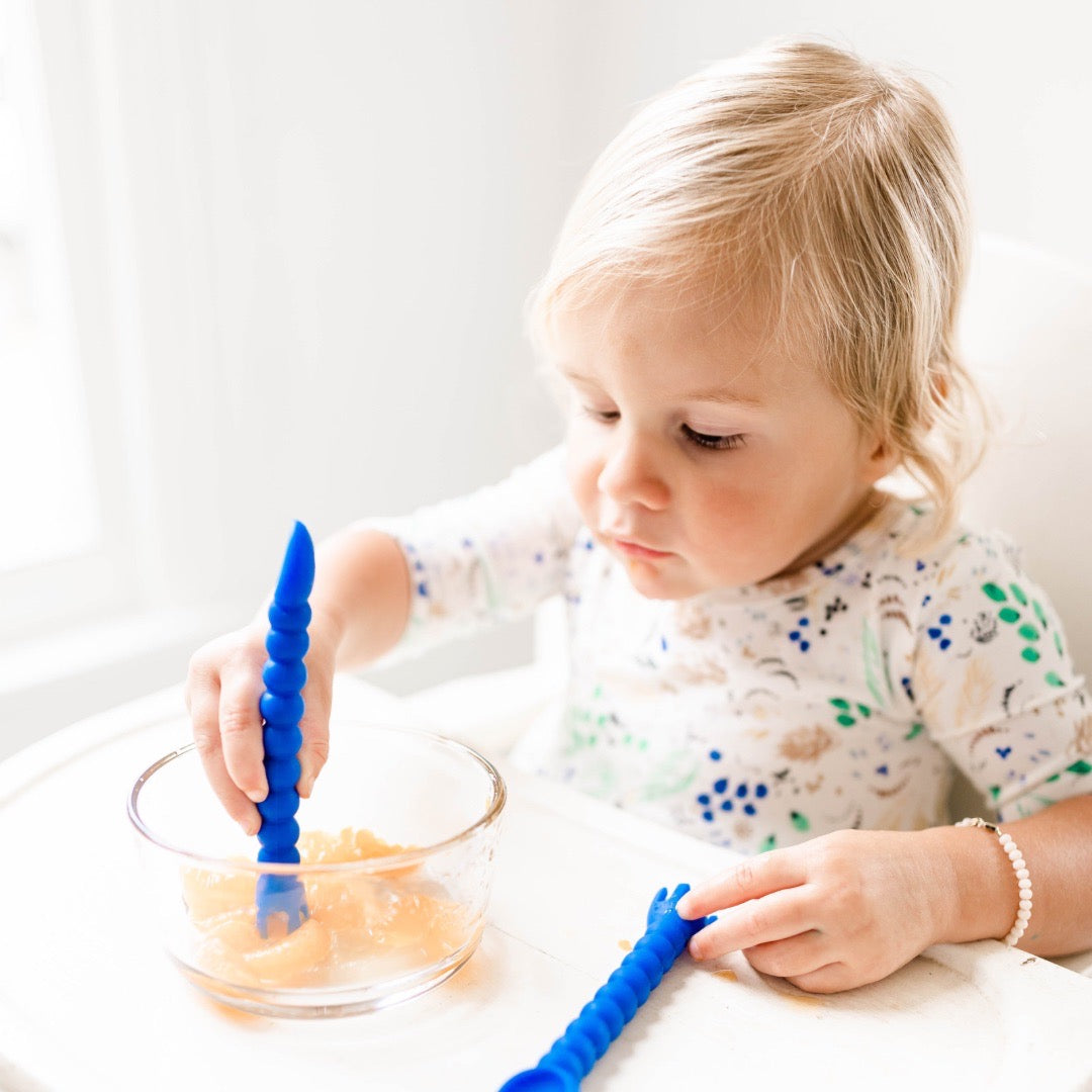 Toddler sitting in high chair eating applesauce using Cutie Tensils.