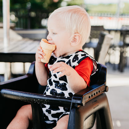 Baby sitting in a high chair wearing the Cow Cutie Bapron and eating an ice cream cone.