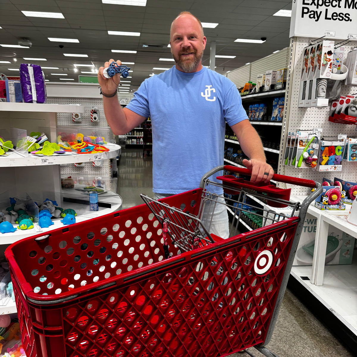 A man in a blue t-shirt stands inside a Target store aisle, holding a small bottle in one hand and pushing a red Target shopping cart with the other hand. Shelves with colorful toys are visible in the background.
