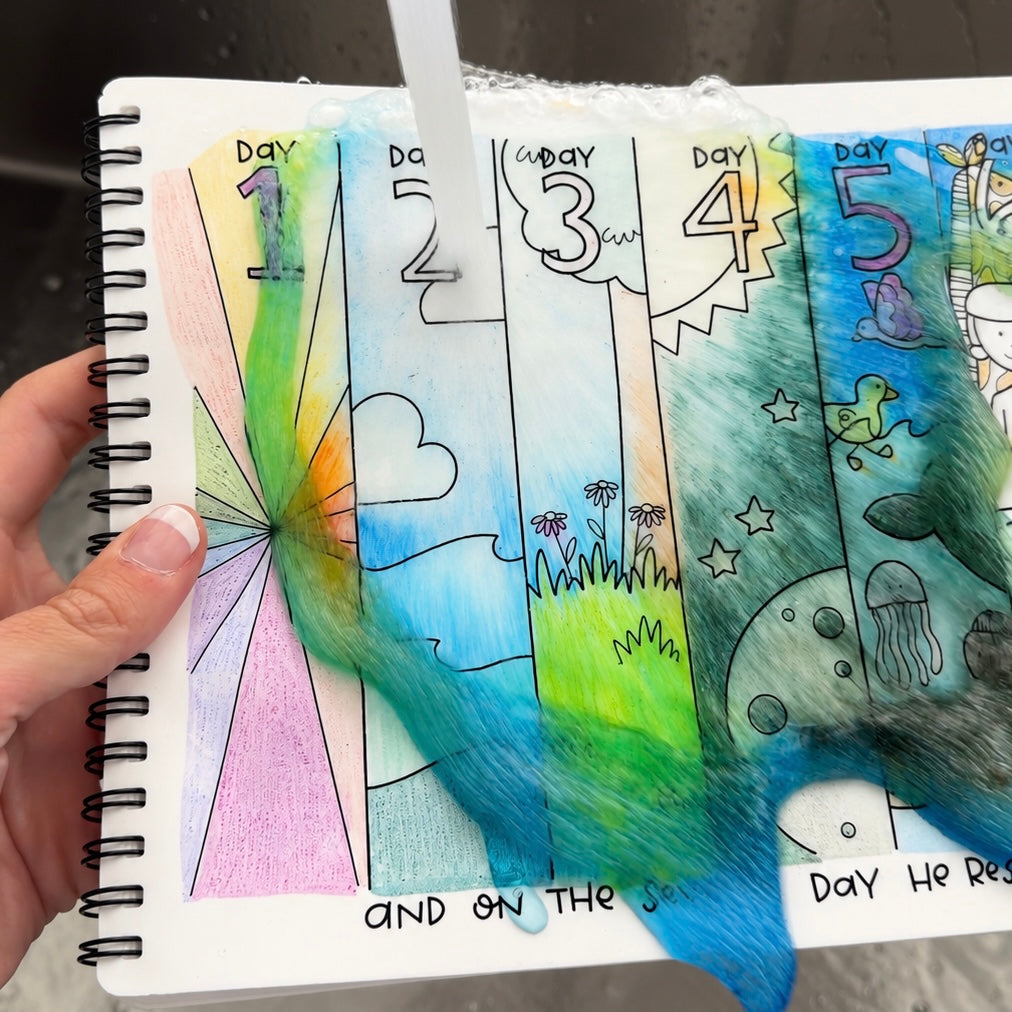 A hand holds a spiral-bound notebook with colorful, water-sensitive daily mood journaling pages. A clear stream of water is running over the pages, causing the colored ink and textured fibers to bleed and shift.