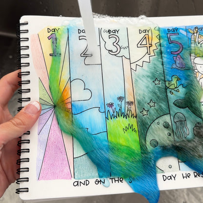 A hand holds a spiral-bound notebook with colorful, water-sensitive daily mood journaling pages. A clear stream of water is running over the pages, causing the colored ink and textured fibers to bleed and shift.