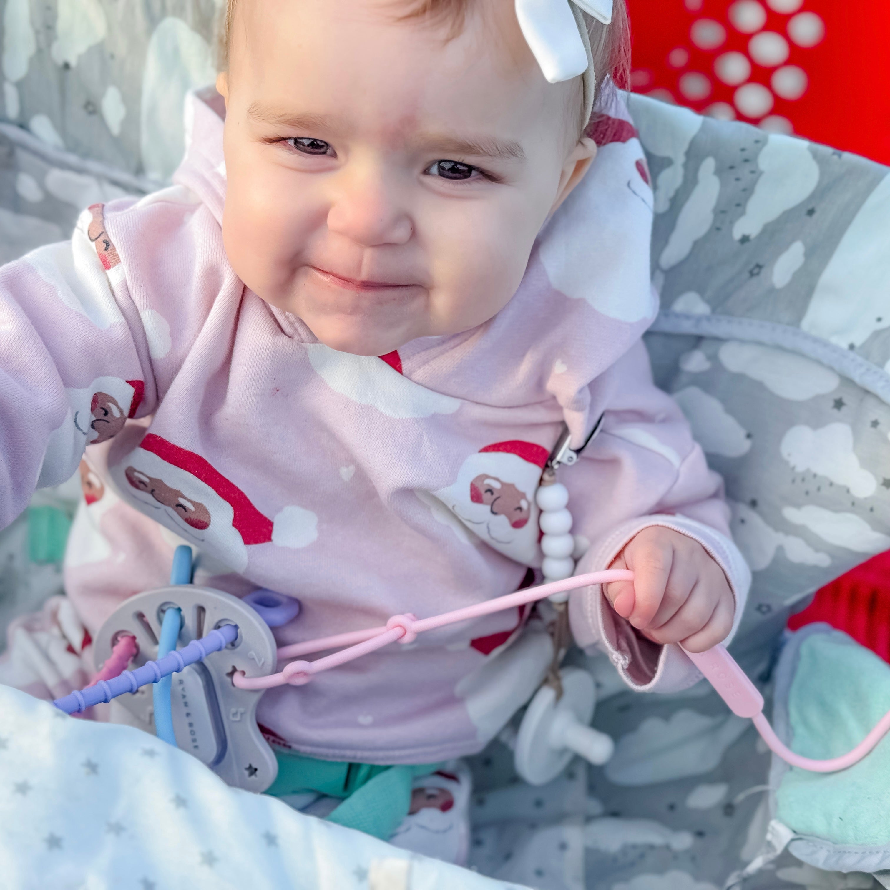 Baby in a pink outfit with a white bow, holding a toy, sitting on a gray and white patterned surface.