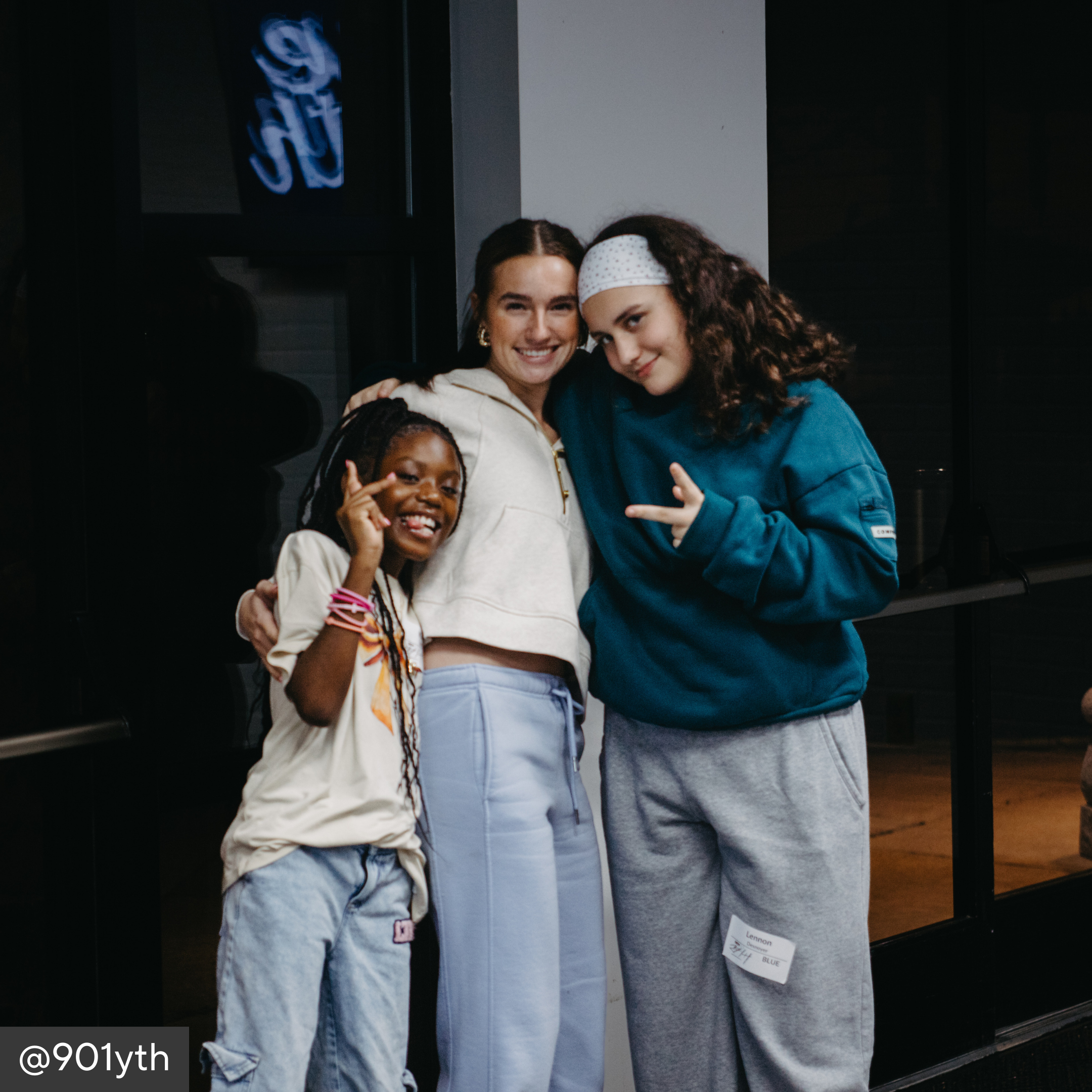 Three people posing together in a casual setting with a neon sign in the background.