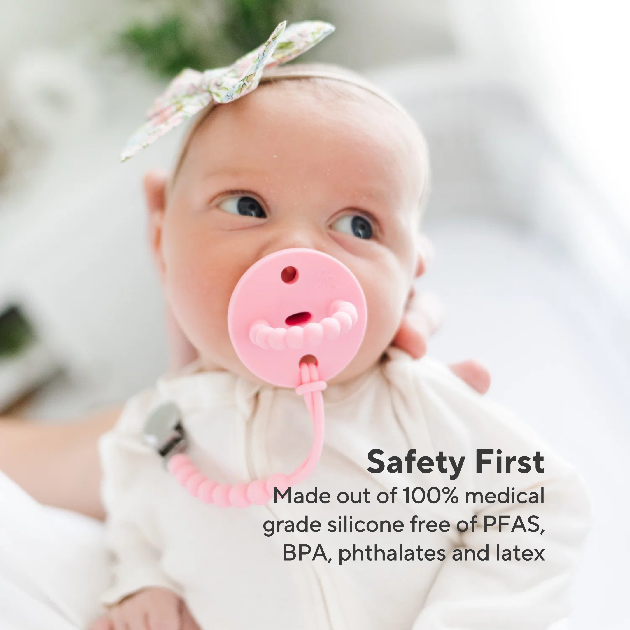 Baby with a pink silicone teether, wearing a white outfit and headband, with a blurred background.