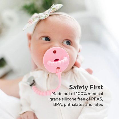Baby with a pink silicone teether, wearing a white outfit and headband, with a blurred background.